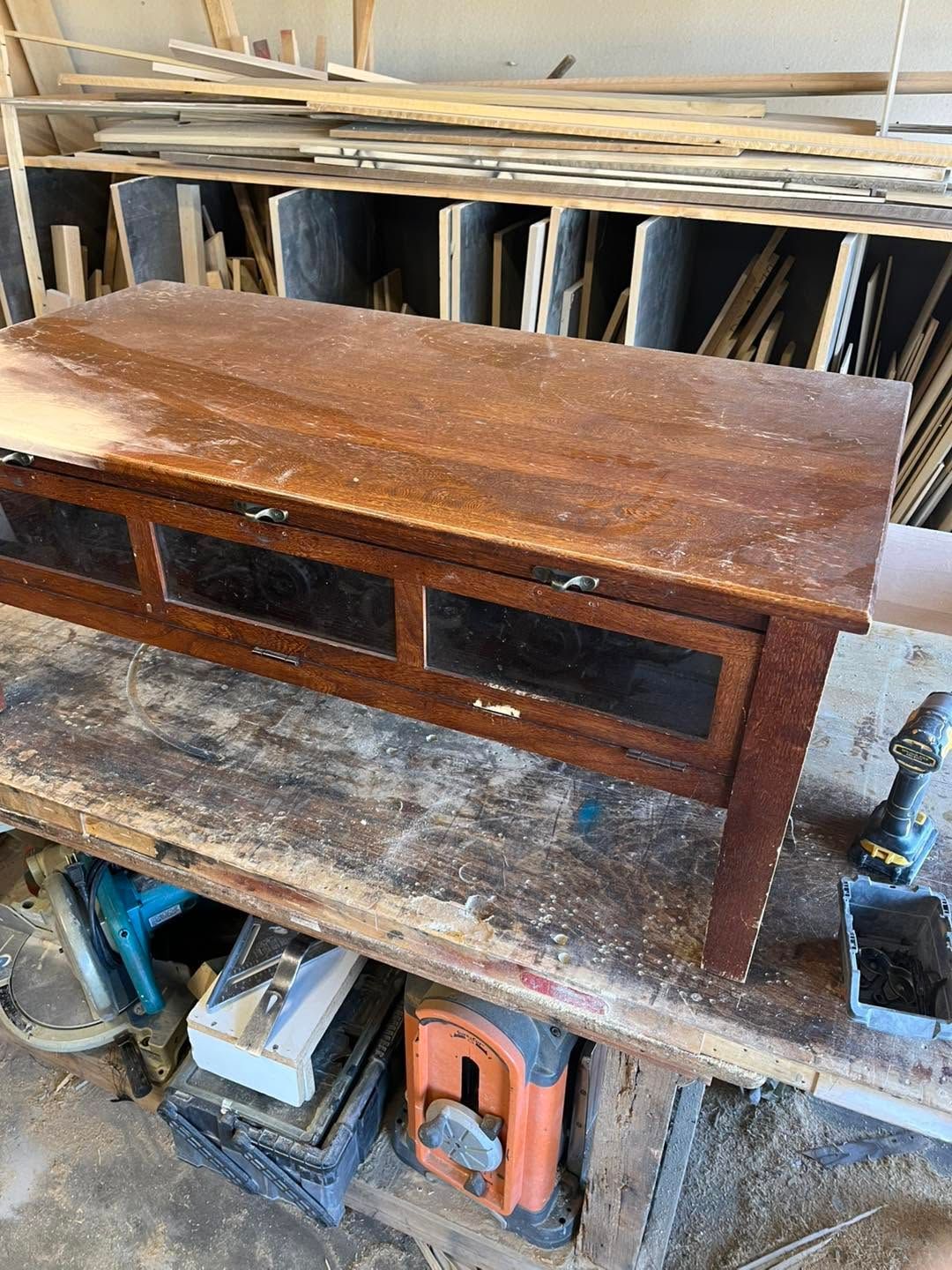Brown wooden cabinet on a workbench in a cluttered workshop, with tools and wood boards in the background