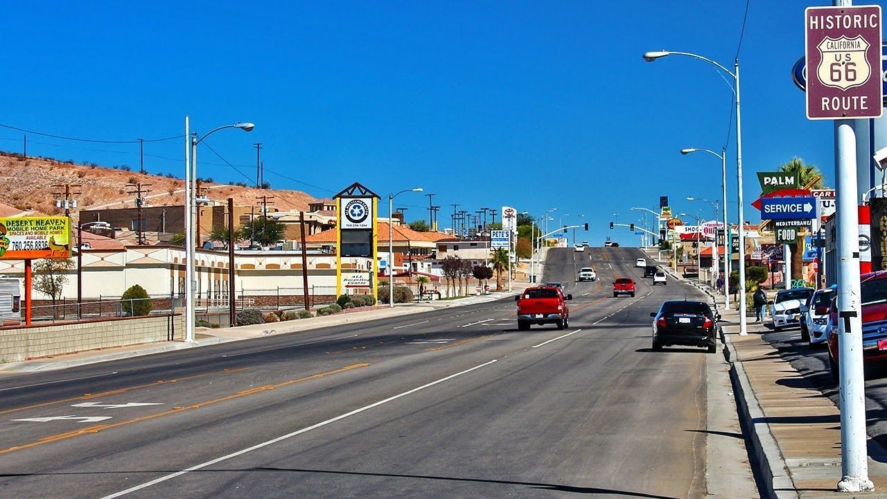 Sunny small-town Route 66 street with cars, storefronts, and desert hills in the background
