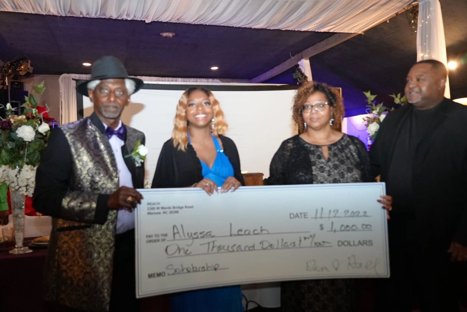Four people holding a large check. Alyssa Leach receives $2,000.  Formal event setting with flowers, a screen, and decorations.