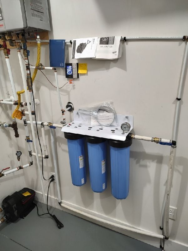 Water filtration equipment with three blue canisters, pipes, valves, and wall-mounted controls in a utility room