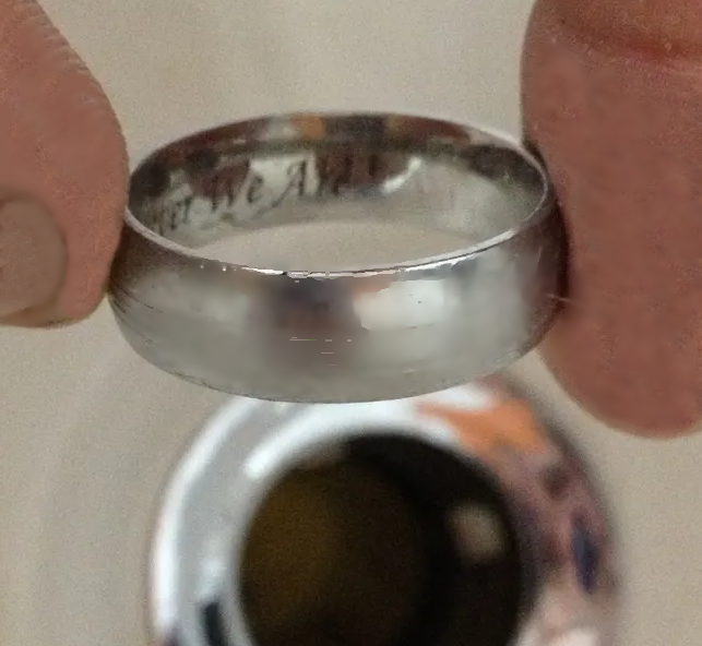 Close-up of a silver ring held between two fingers, with a blurred sink in the background.