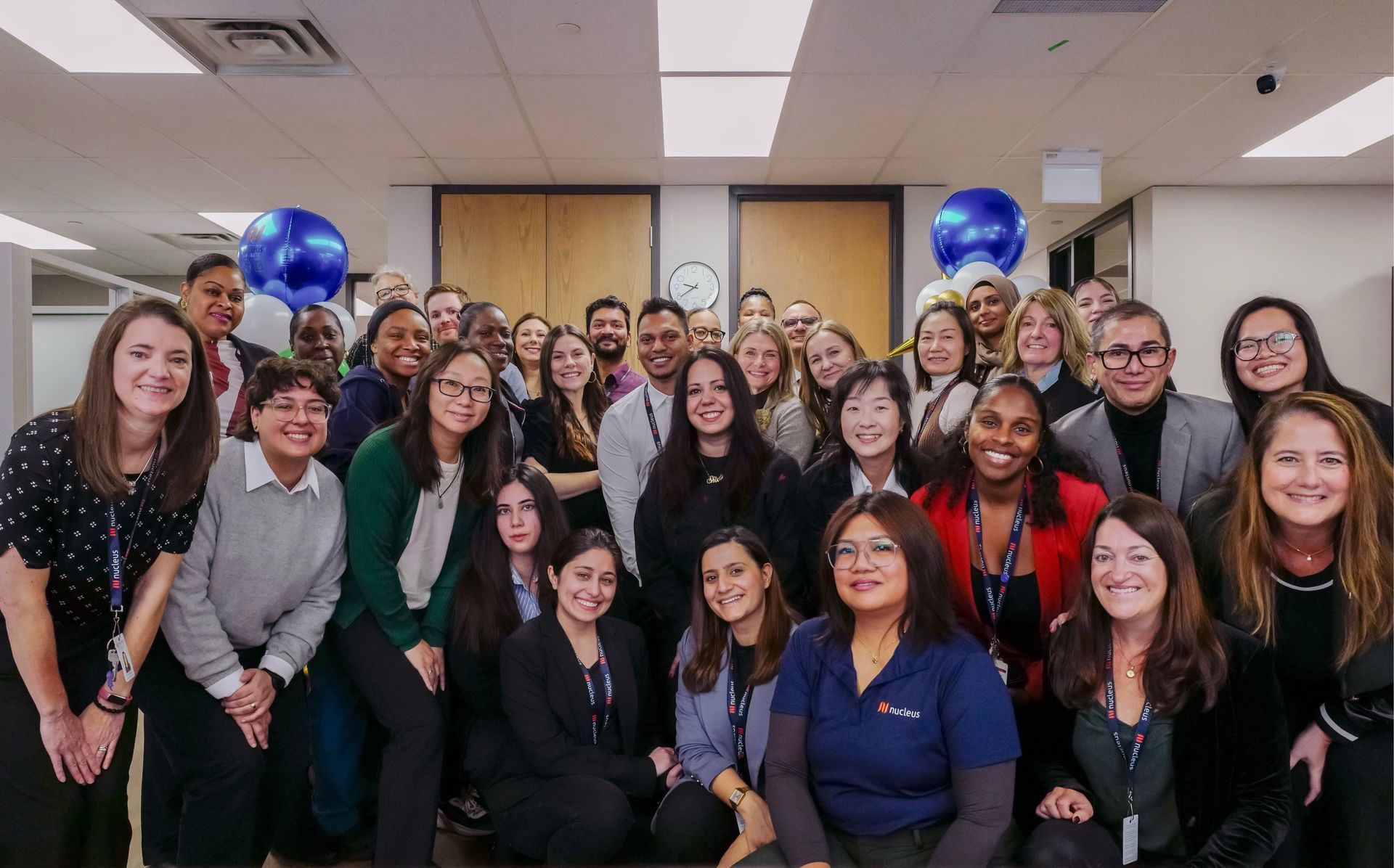 Group of smiling colleagues posing in an office, with blue balloons and conference room doors in the background