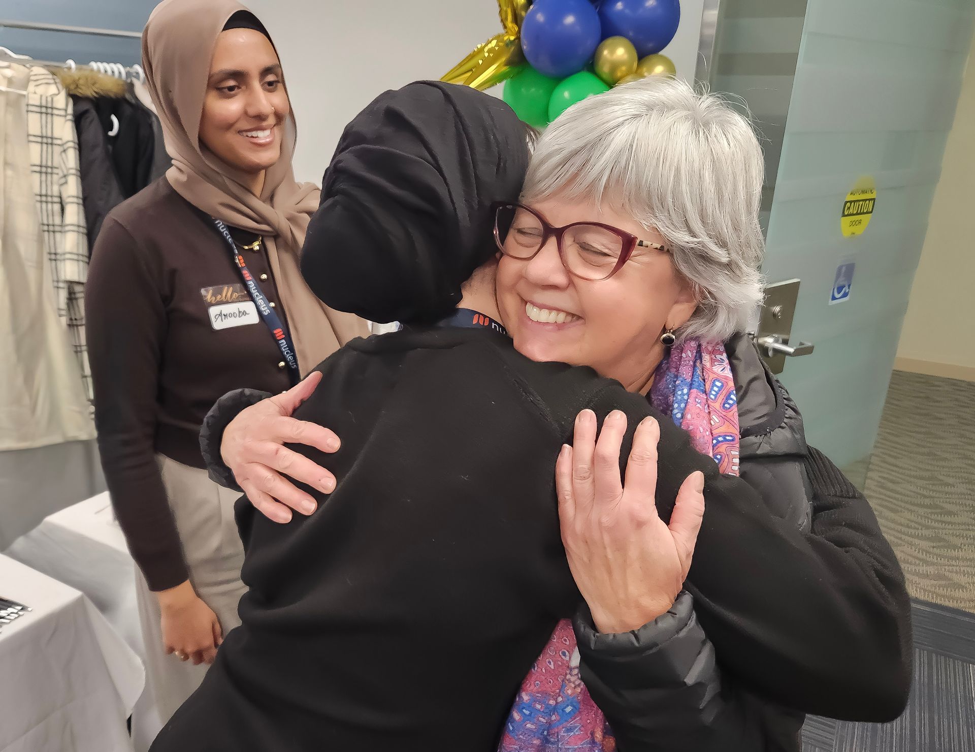 Two people hugging in a bright indoor setting, with balloons in the background and a smiling woman nearby