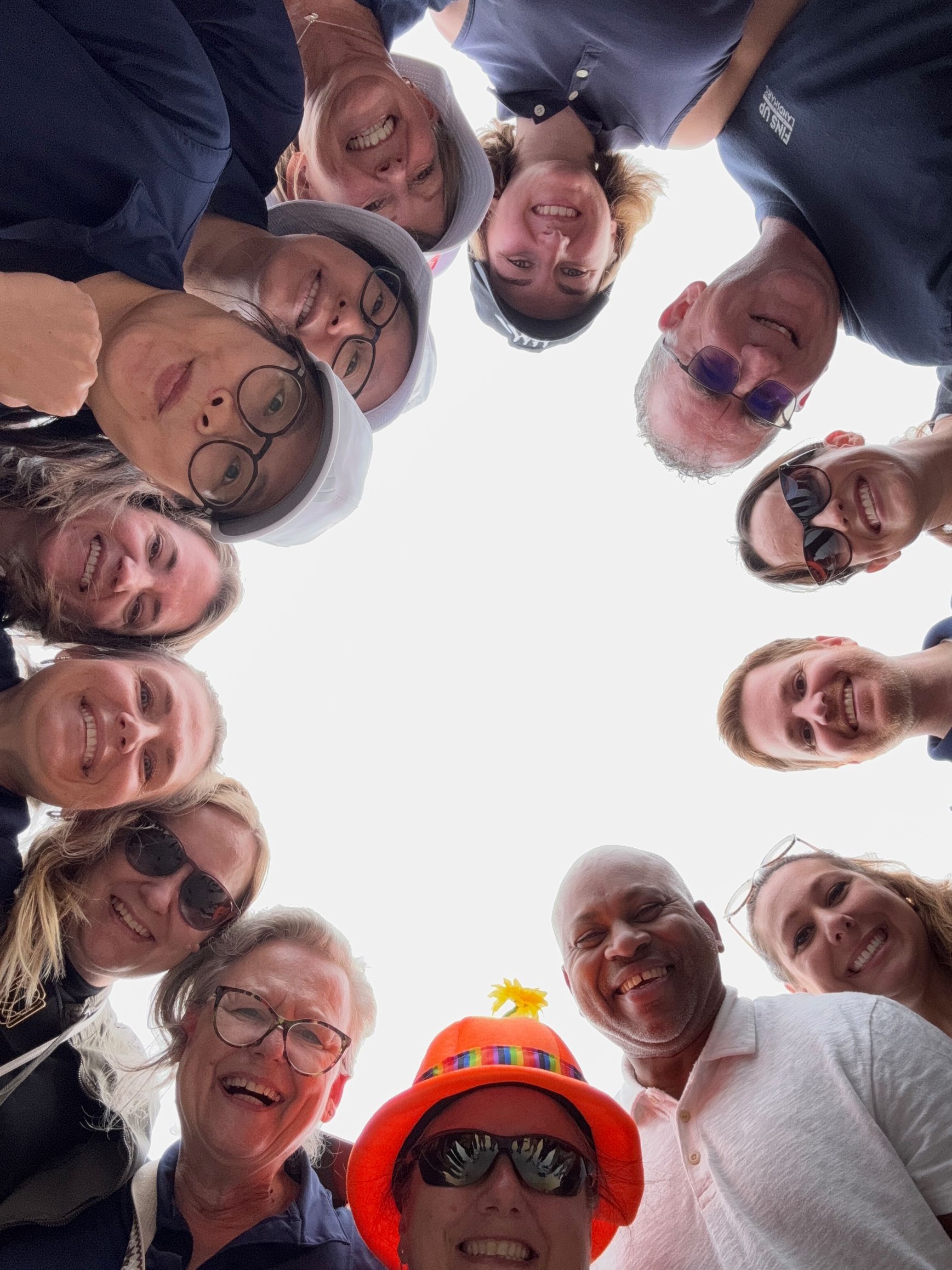 Group of people leaning in and smiling around the camera, with one person in a bright orange hat and sunglasses.