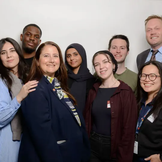 Seven colleagues smiling and posing together in a studio against a light gray background