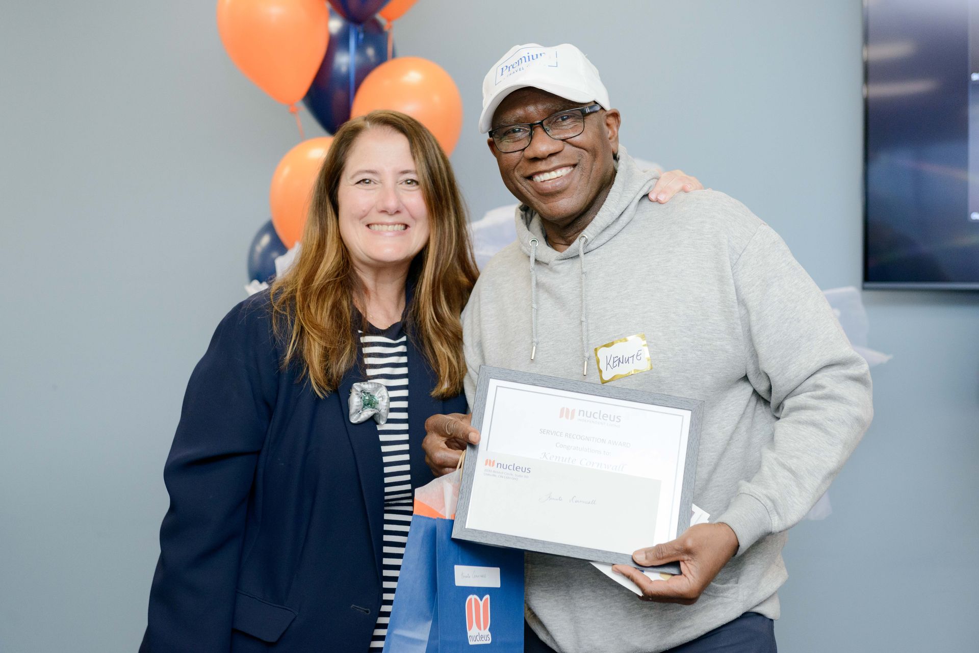 Two smiling people holding a certificate at an event, with orange balloons in the background.