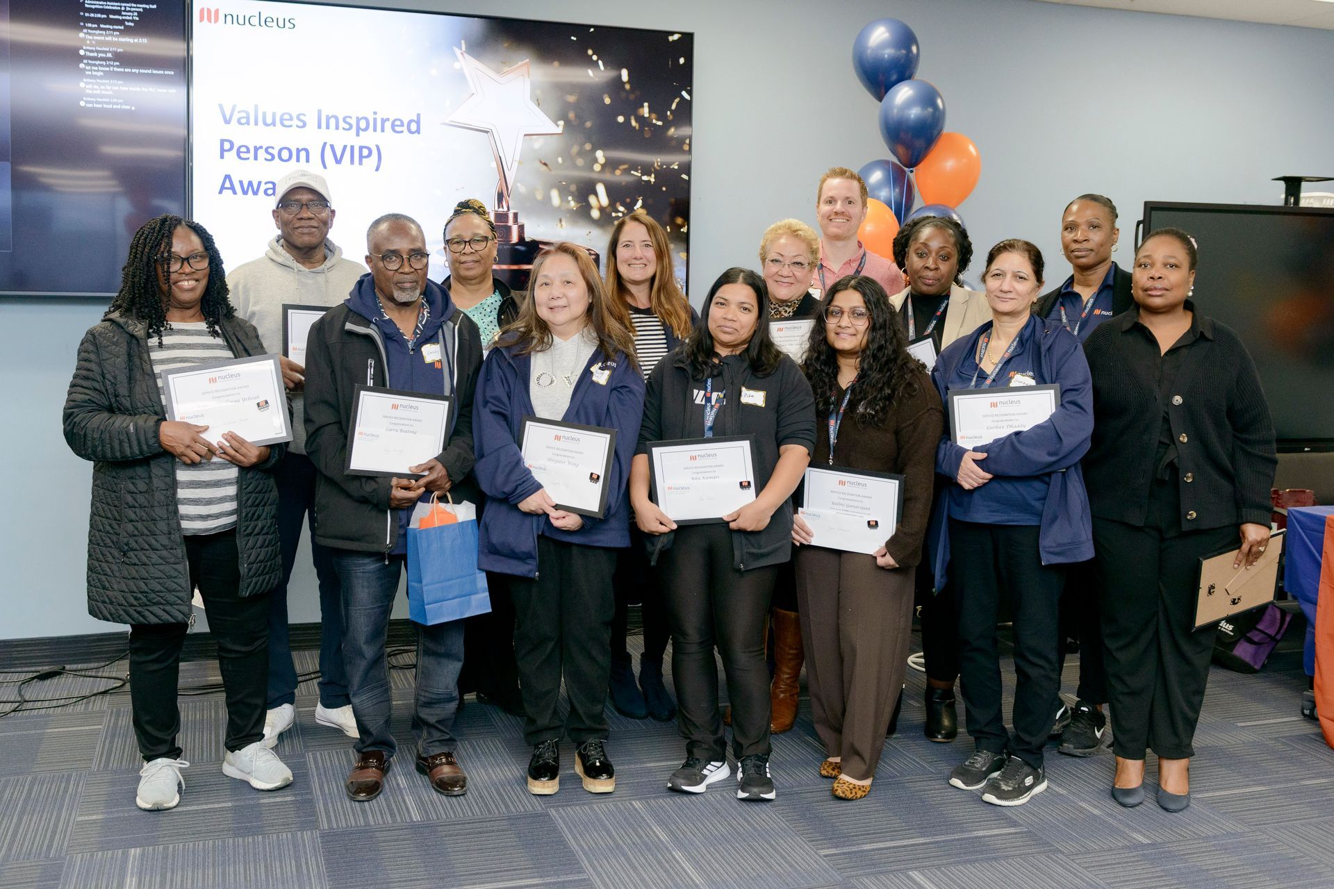 Group of people holding certificates at a workplace celebration with blue and orange balloons