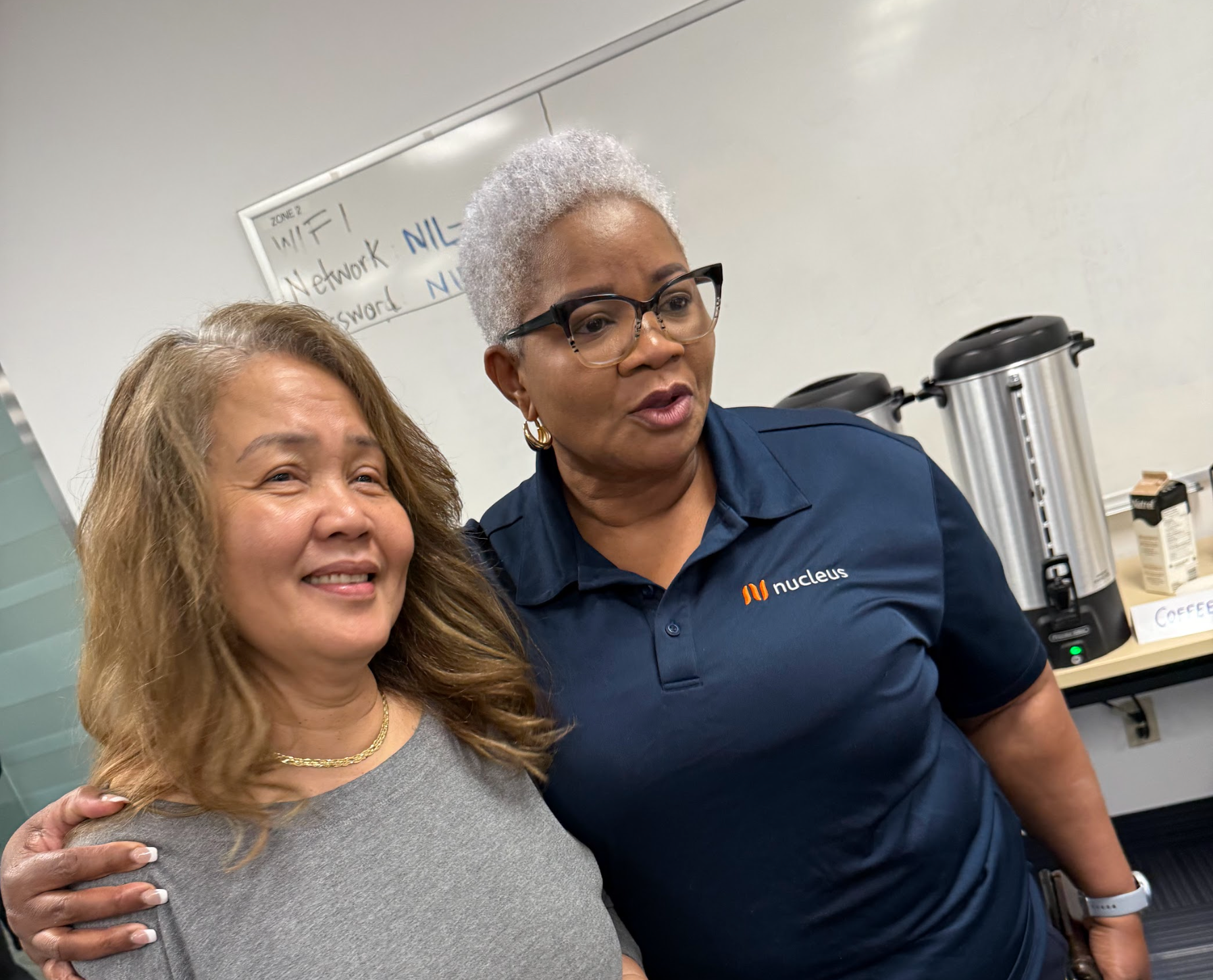 Two women smiling beside a coffee machine in an office break room