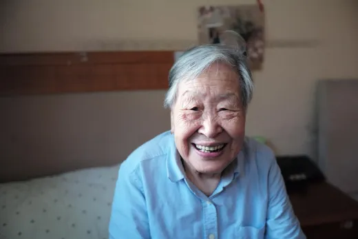 Smiling elderly woman in a light blue shirt sitting indoors on a bed