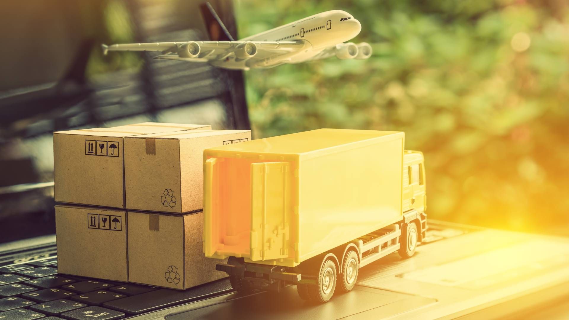 A toy truck is sitting on top of a laptop with boxes and an airplane flying in the background.