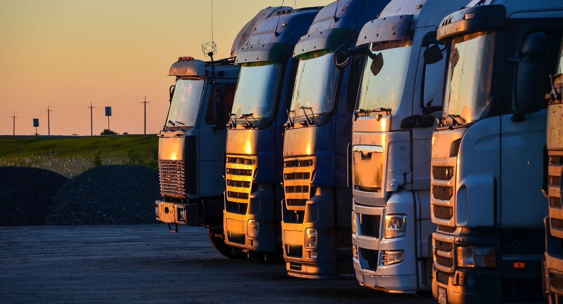 A row of semi trucks are parked in a parking lot at sunset.