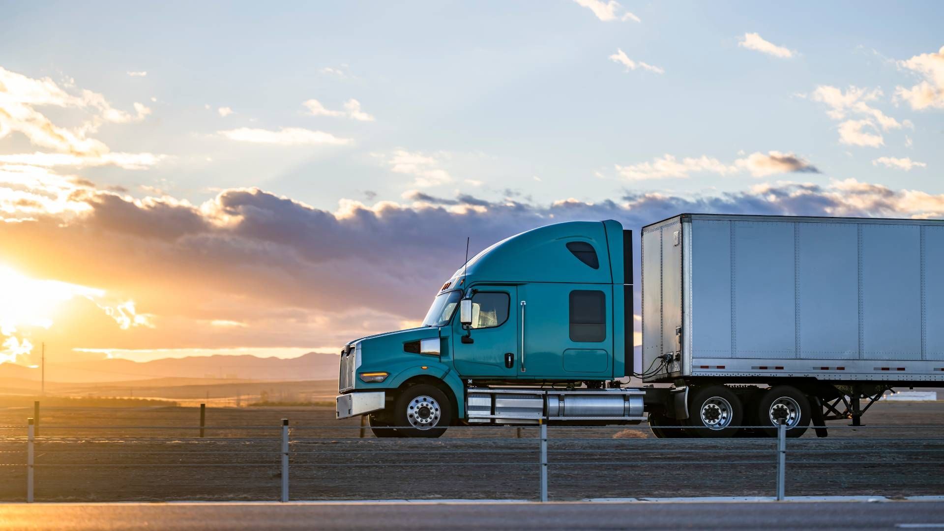 A blue semi truck is driving down a highway at sunset.