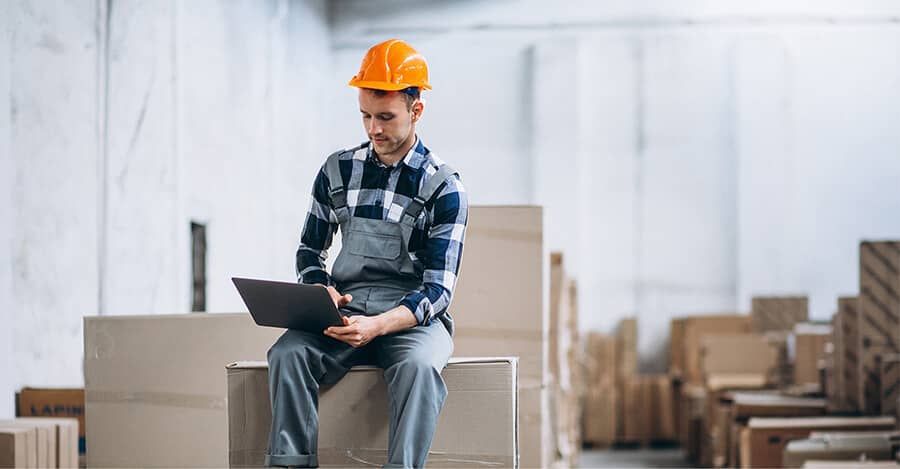 A man is sitting on a pile of cardboard boxes in a warehouse using a laptop.