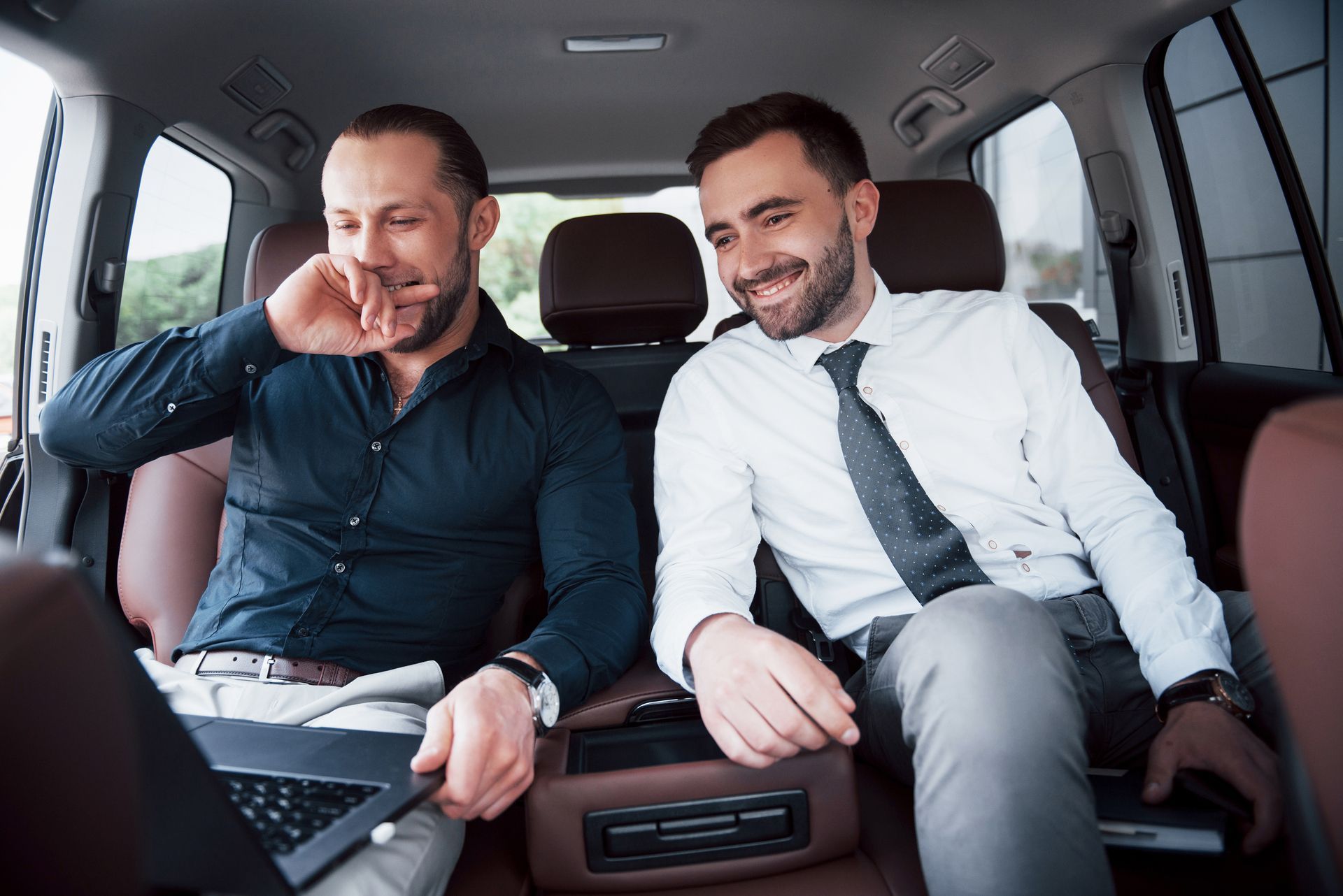 Two men are sitting in the back seat of a car looking at a laptop.