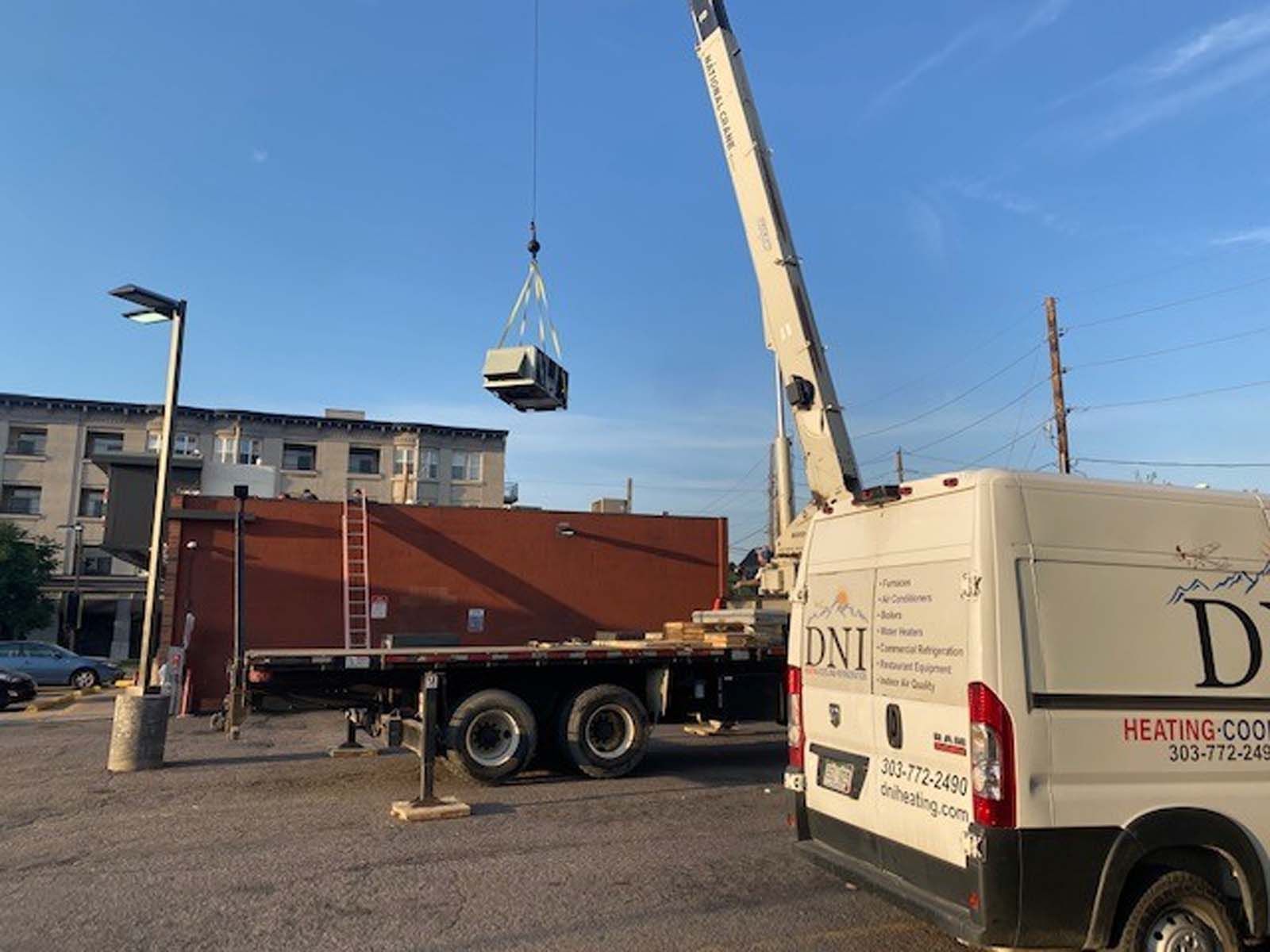 A crane lifts an HVAC unit over a flatbed truck in a parking lot. A building and D.N.I. Heating and Cooling van are in view.