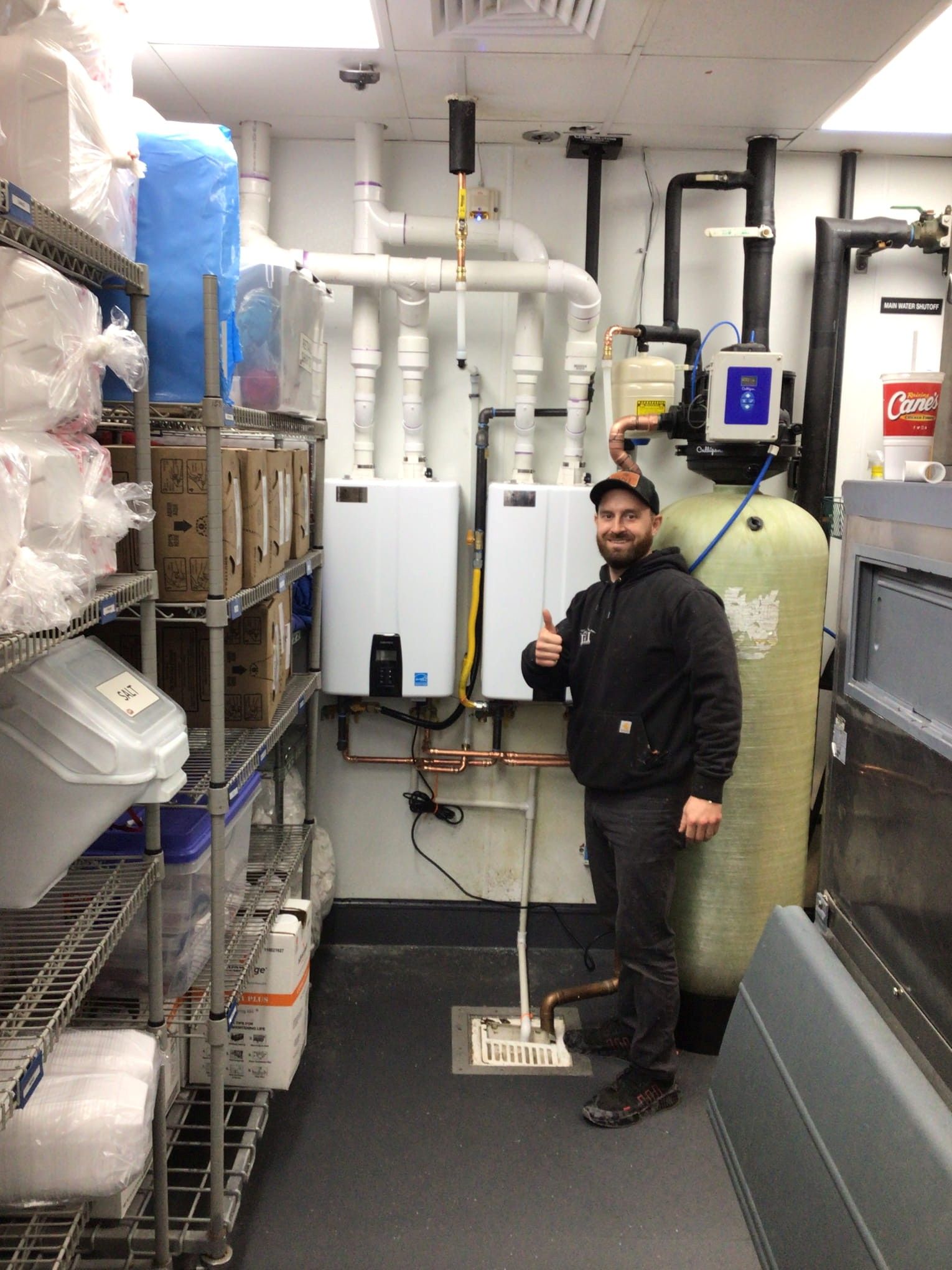 Man giving thumbs up next to water heaters in a utility room. Shelves with supplies on the left.