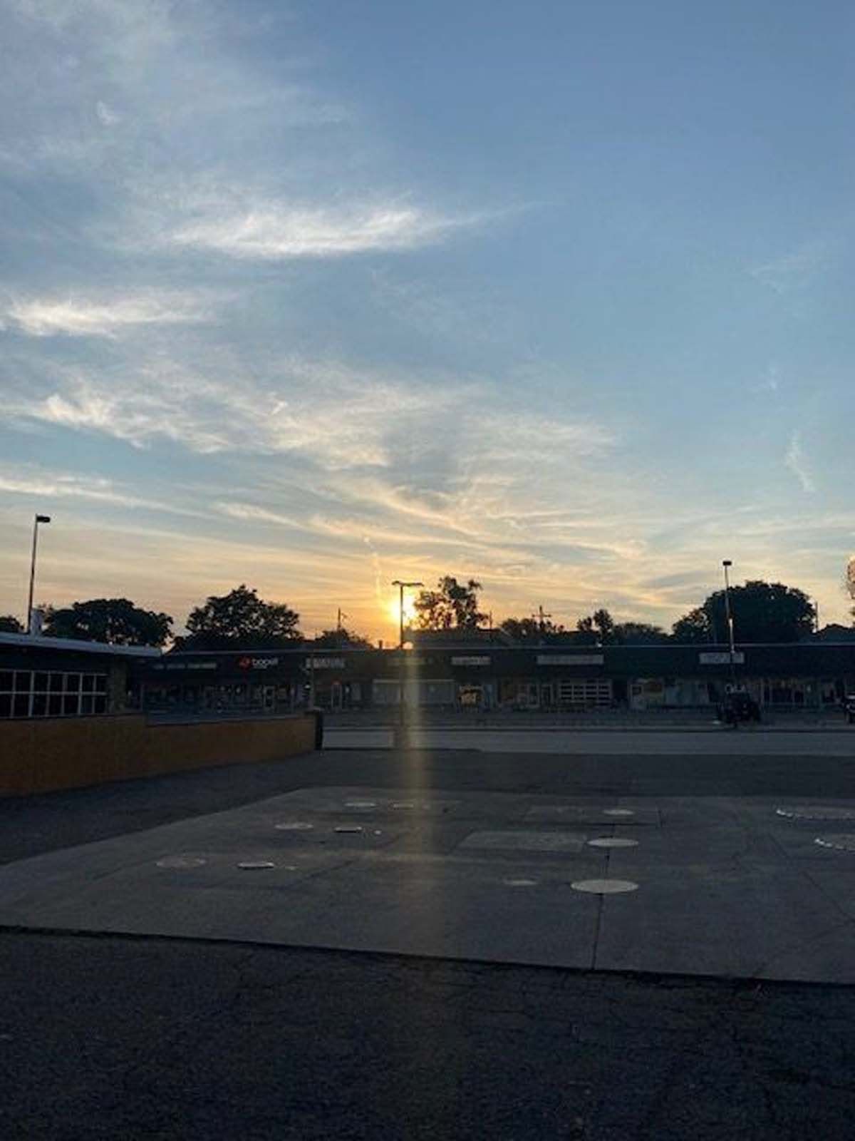 Sunrise over a bus station; light casts a glare, with trees and building silhouettes. The sky is blue with wispy clouds.
