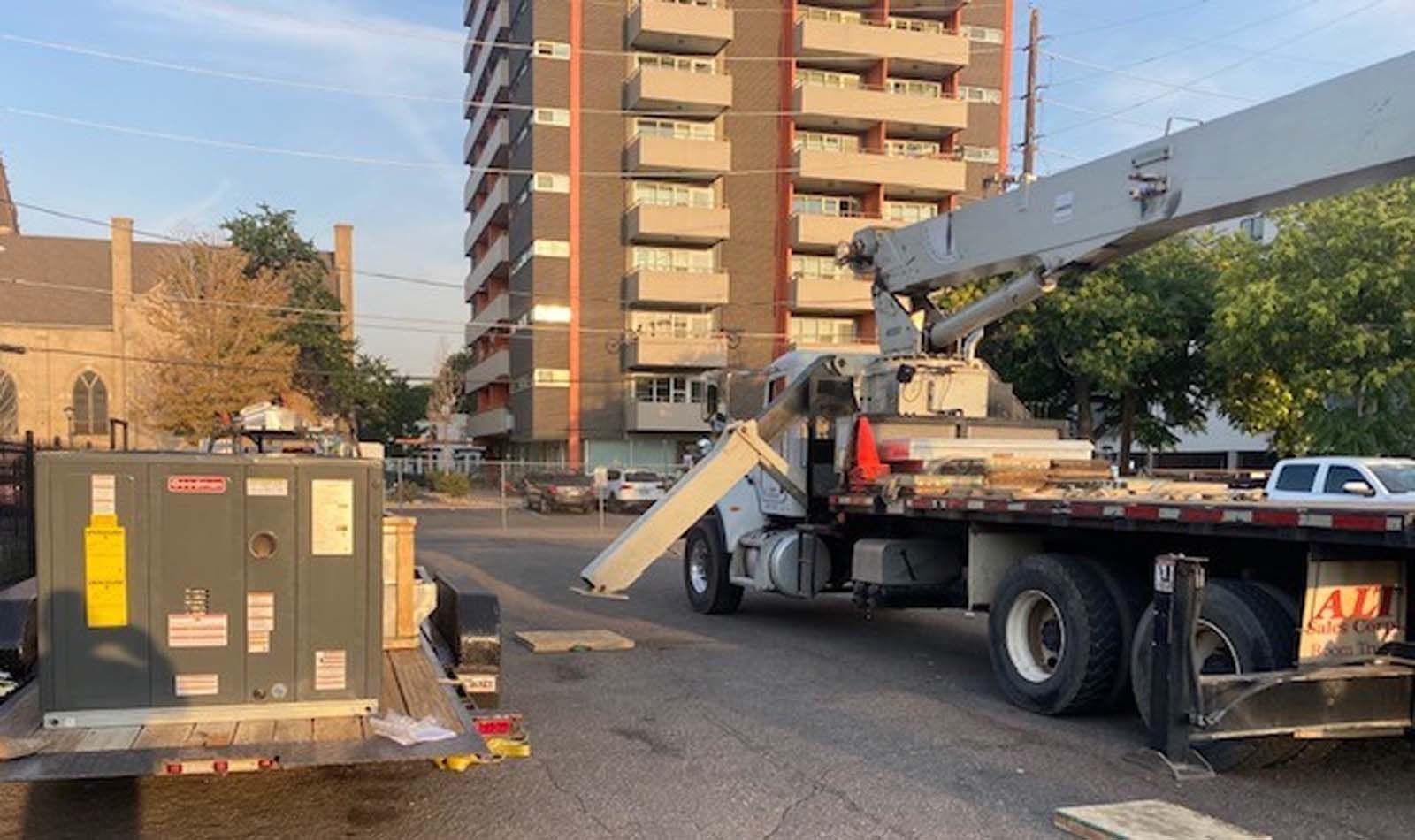 A crane truck is parked on a street with a tall apartment building in the background. A trailer with equipment is on the left.