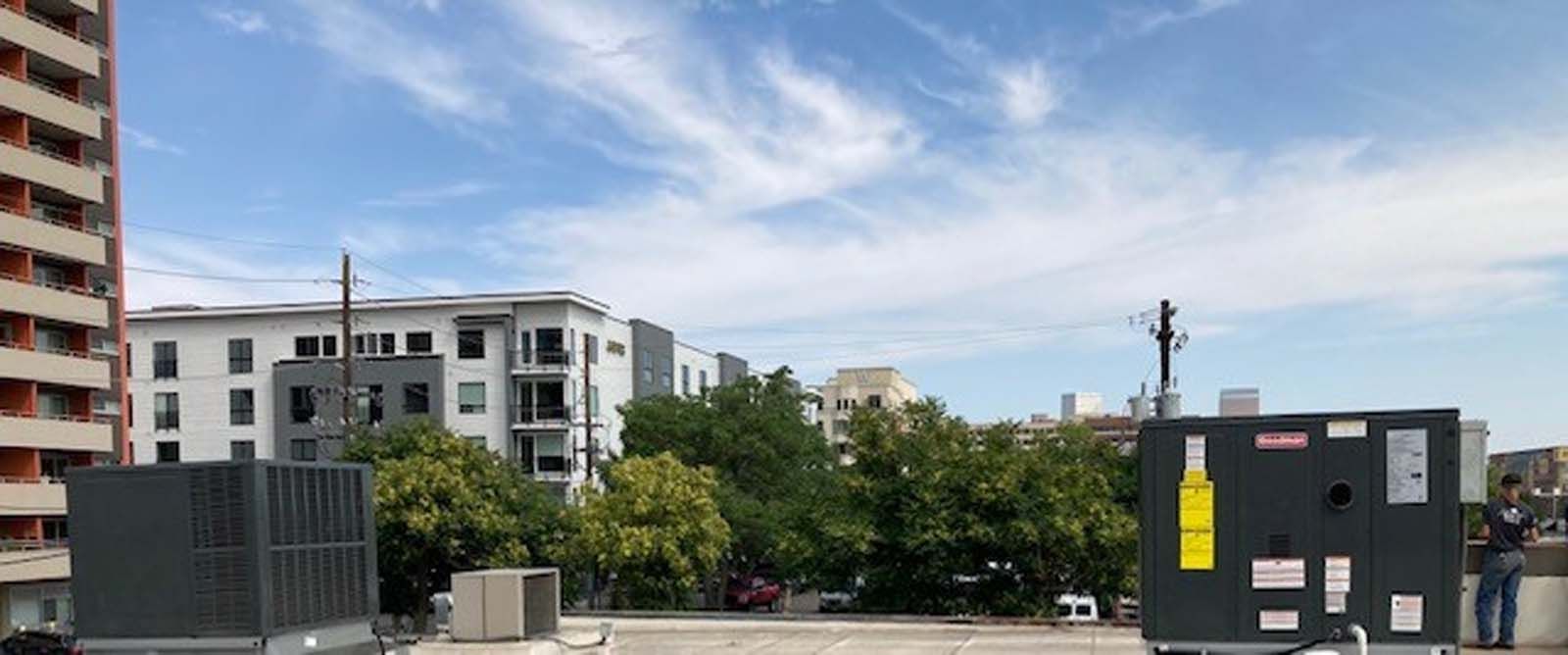 Rooftop view of city buildings under a blue sky with wispy clouds. Large HVAC units and trees are in the foreground.