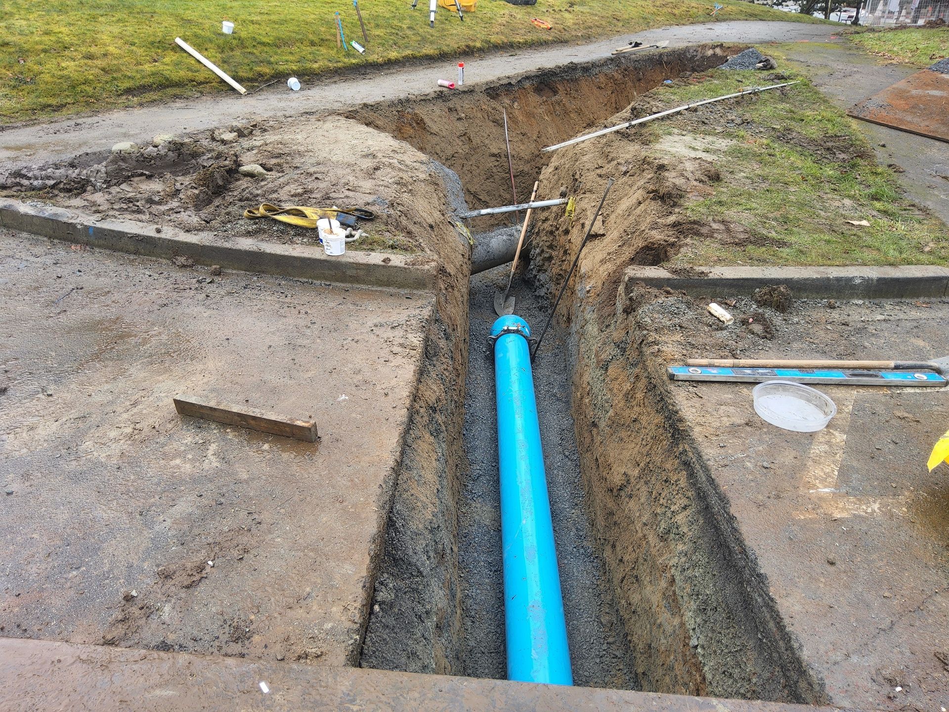 Construction trench with blue pipe laid in it. Concrete path, grass, and tools surround the site.