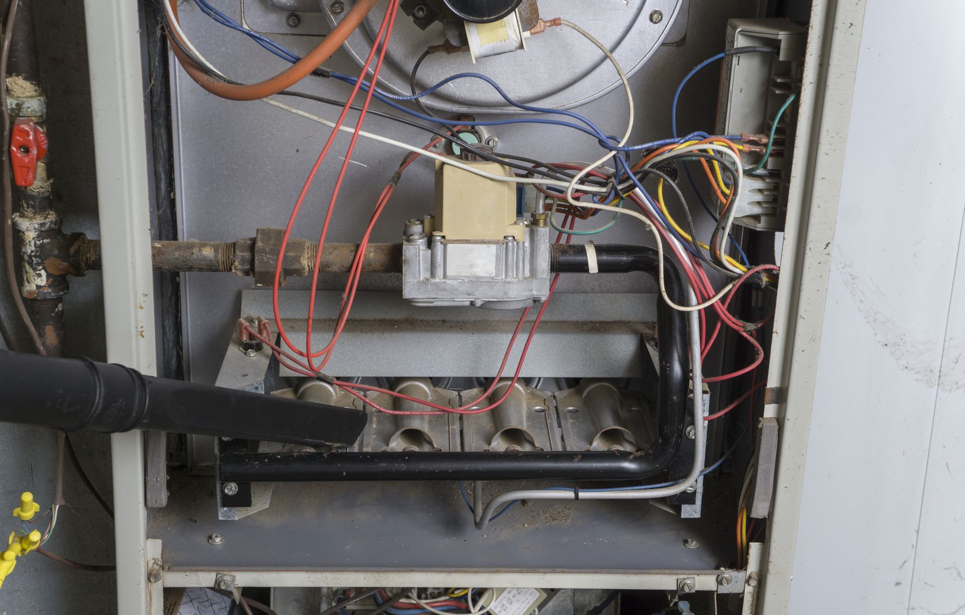 Open furnace interior being cleaned with a black vacuum nozzle, showing wires, pipes, and gas burner assembly.