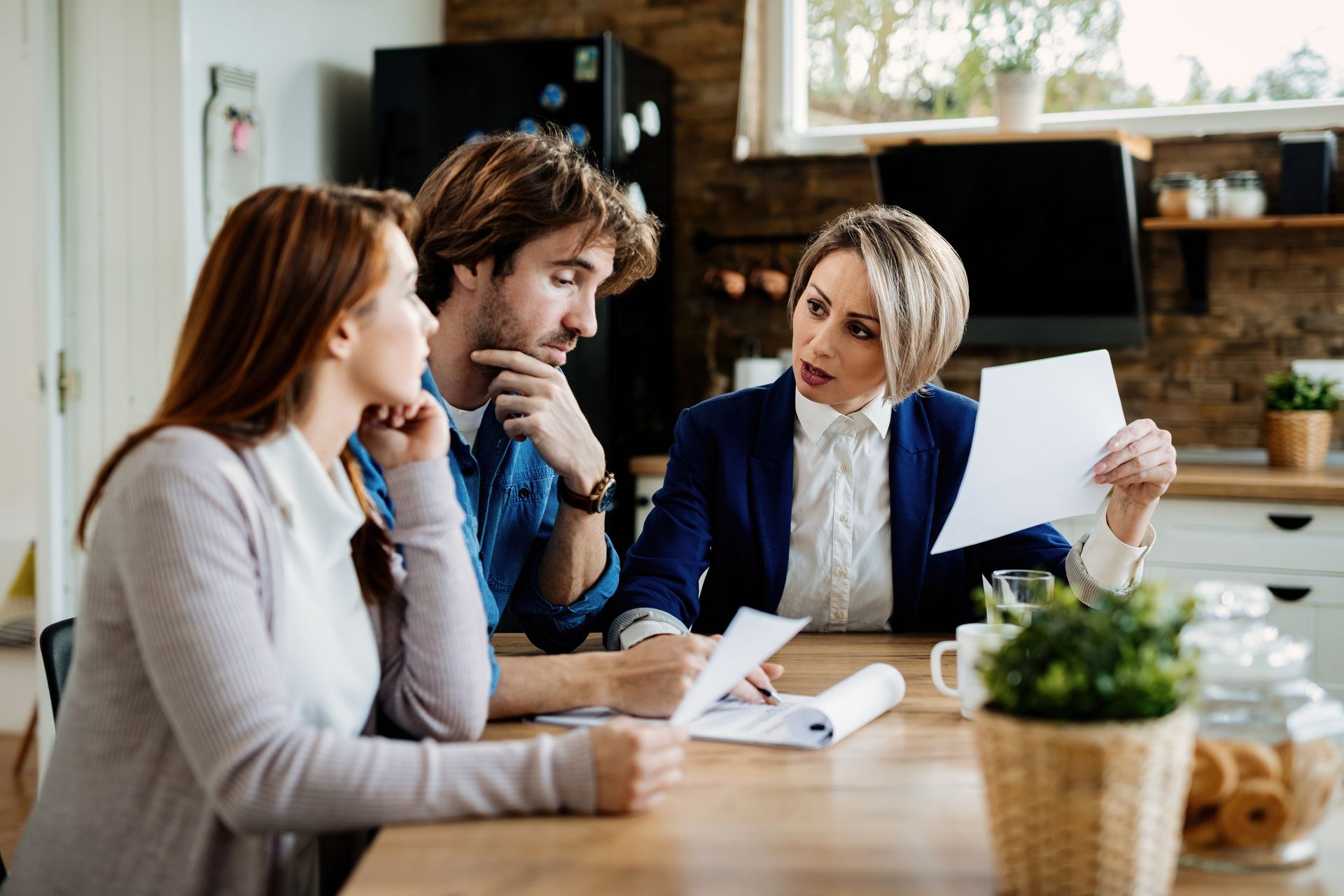 Three coworkers discuss documents at a table in a bright office, one holding a sheet of paper.