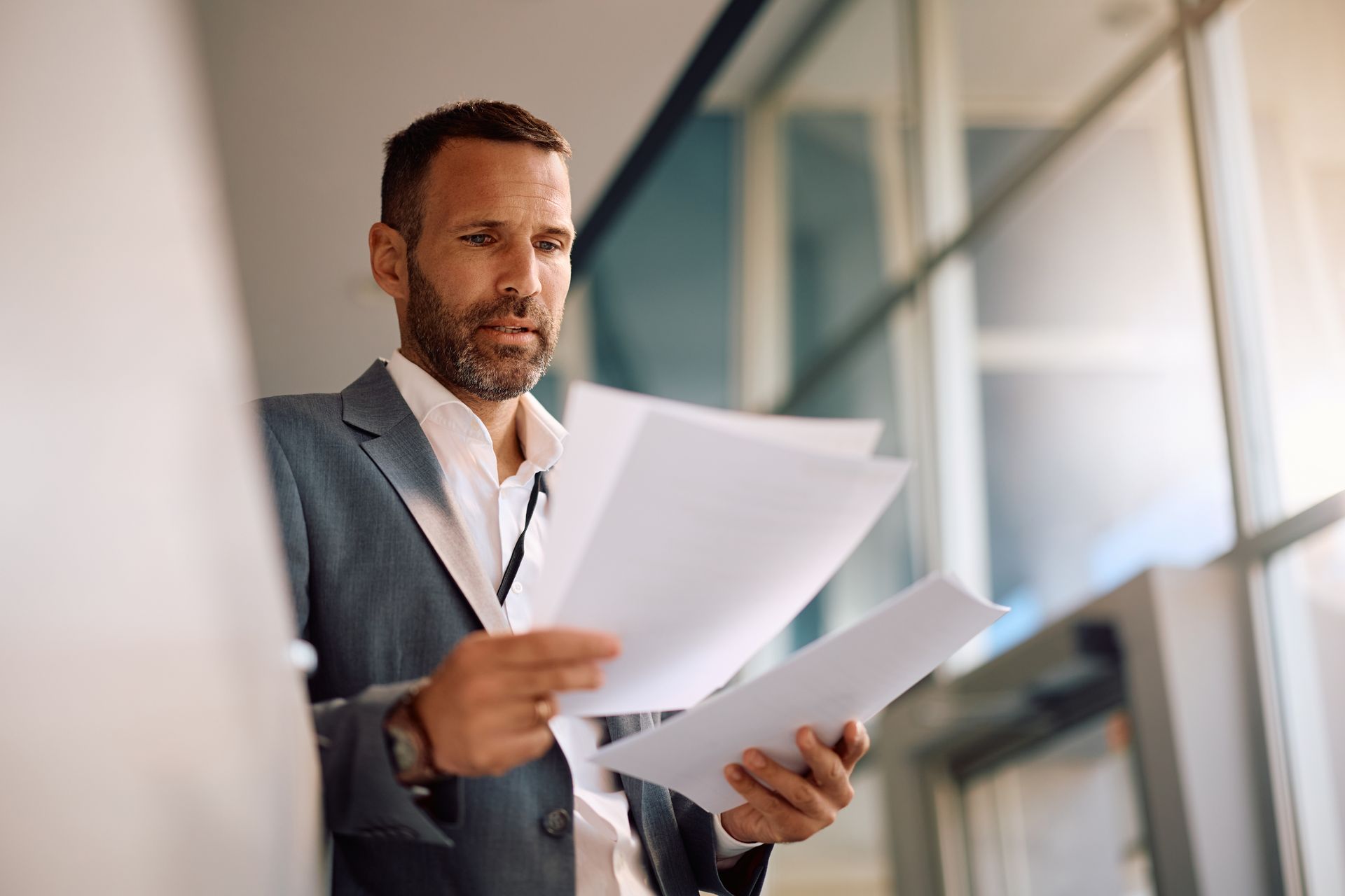 Man in a gray blazer reviewing papers in a bright office hallway