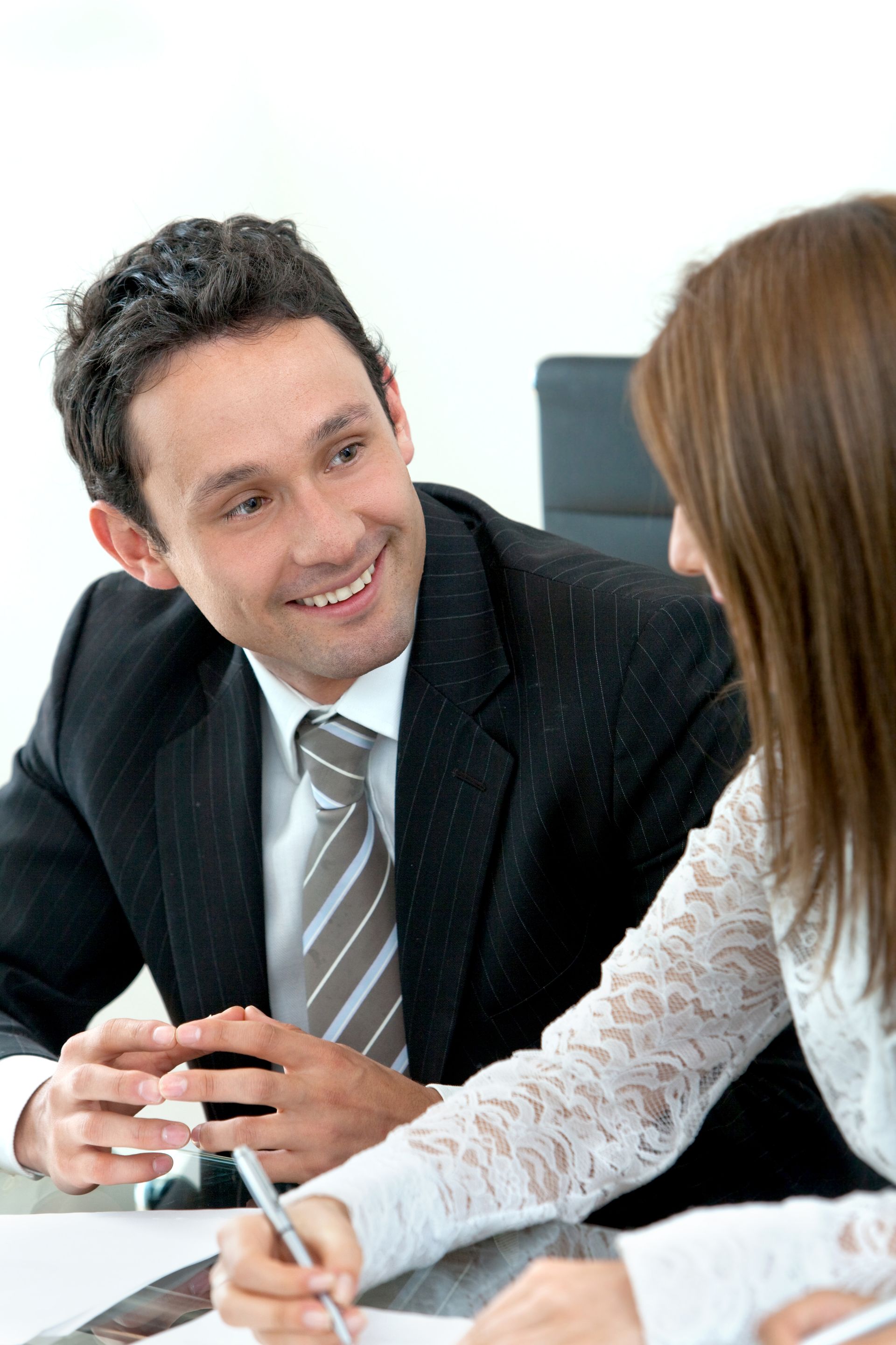 Two coworkers in a meeting, smiling and talking across a table with a notebook and pen.
