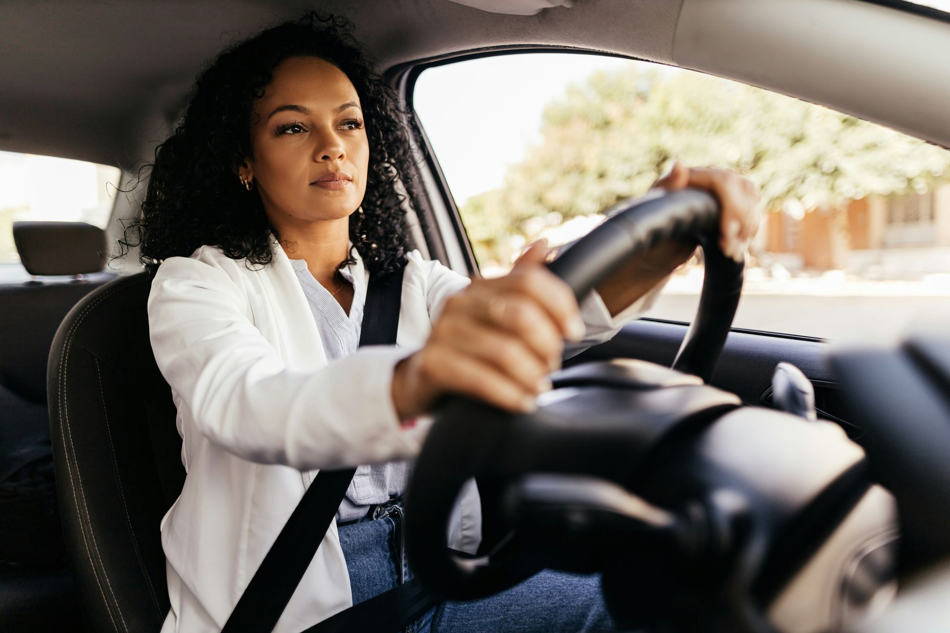 Woman driving a car, hands on the steering wheel, wearing a white shirt and seatbelt.