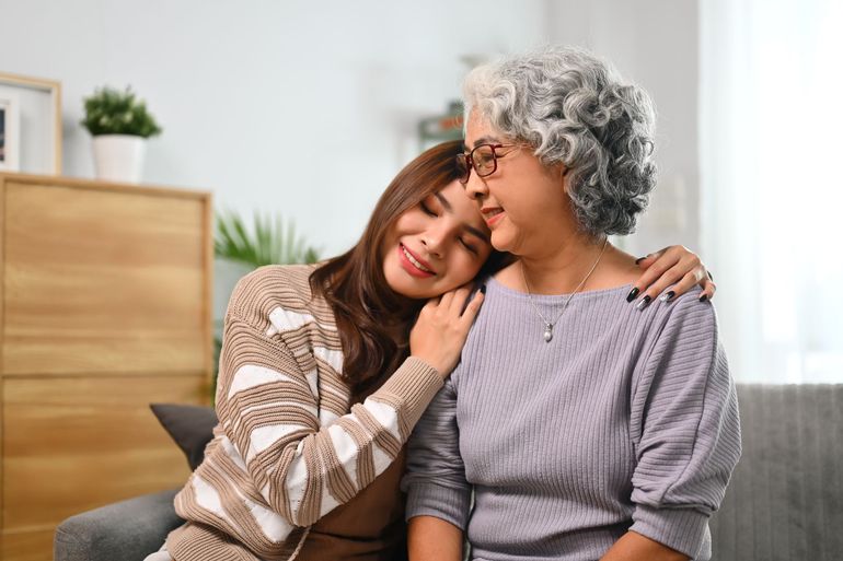 Two women sitting on a couch, one resting her head on the other's shoulder in a cozy living room.