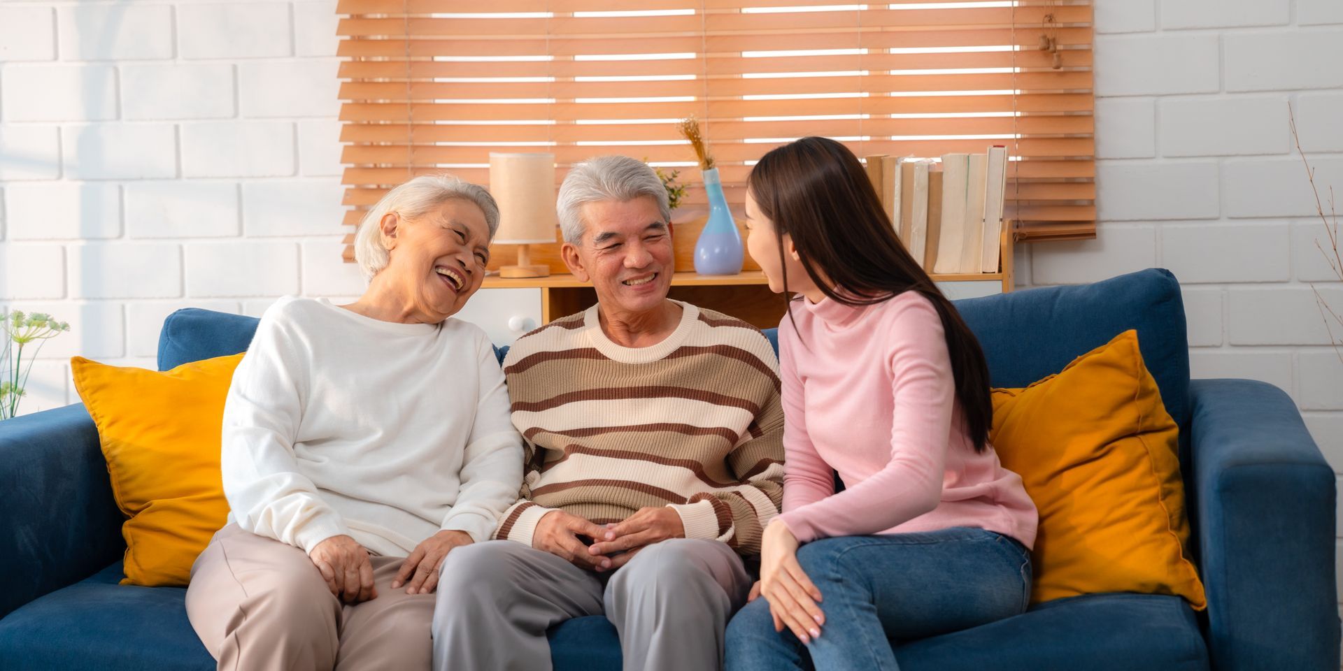 Three people sit on a blue couch, smiling and talking in a bright living room with yellow pillows.