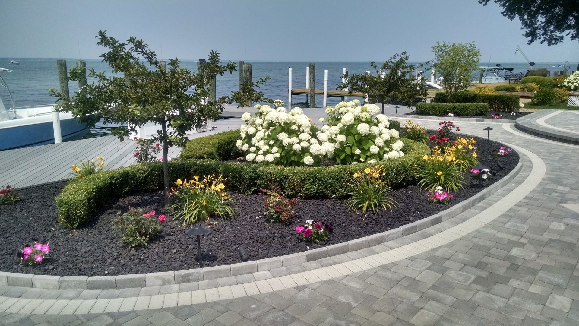 A circular garden with flowers and bushes in front of a body of water
