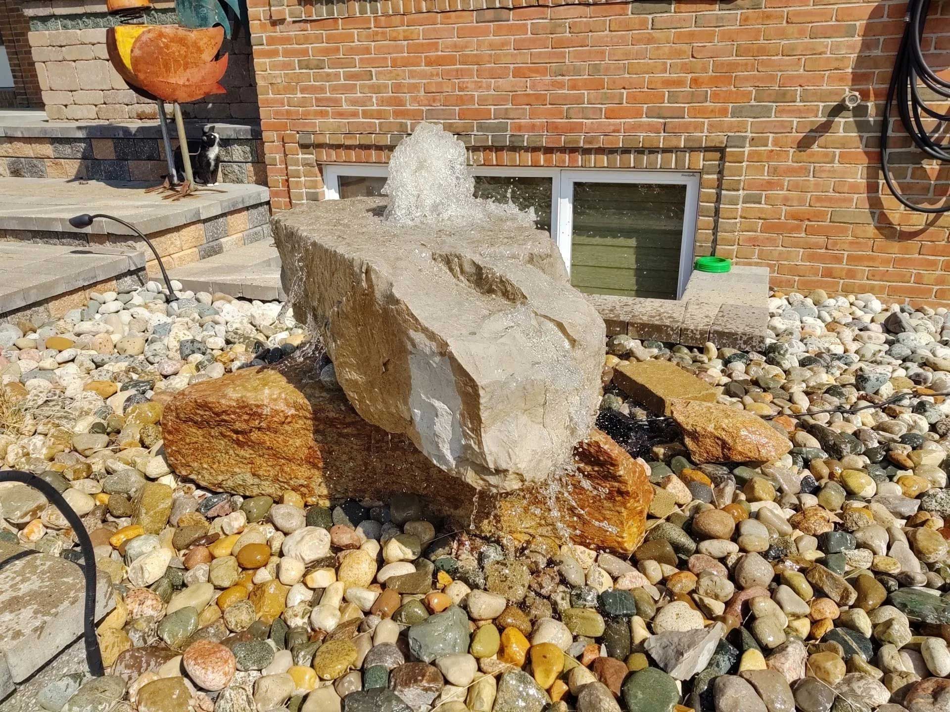 A fountain is sitting on top of a pile of rocks in front of a brick building.