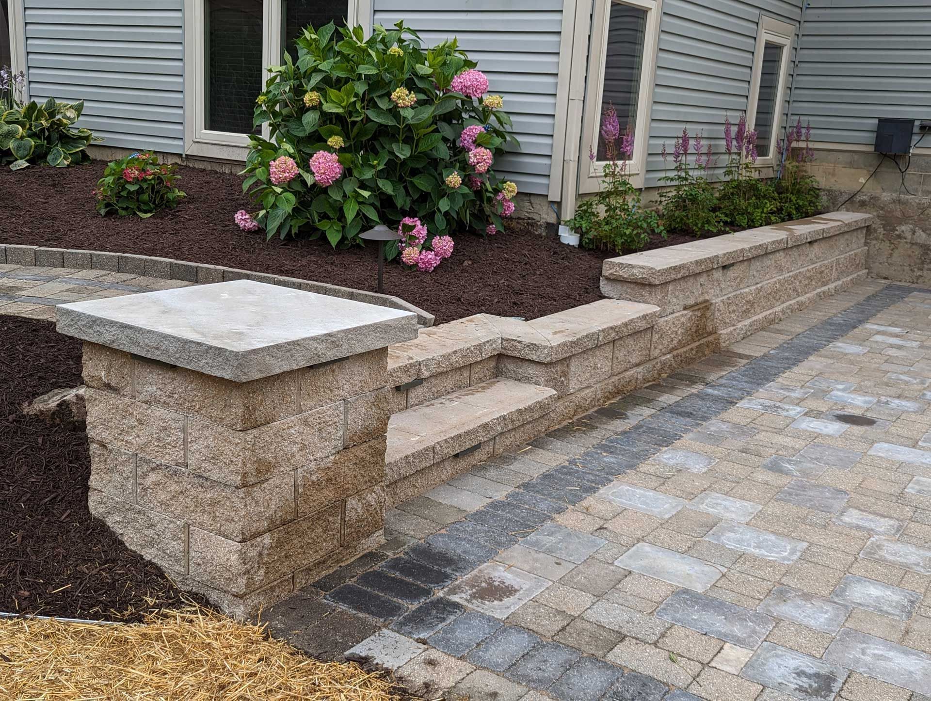 A brick walkway leading to a house with flowers in the background.