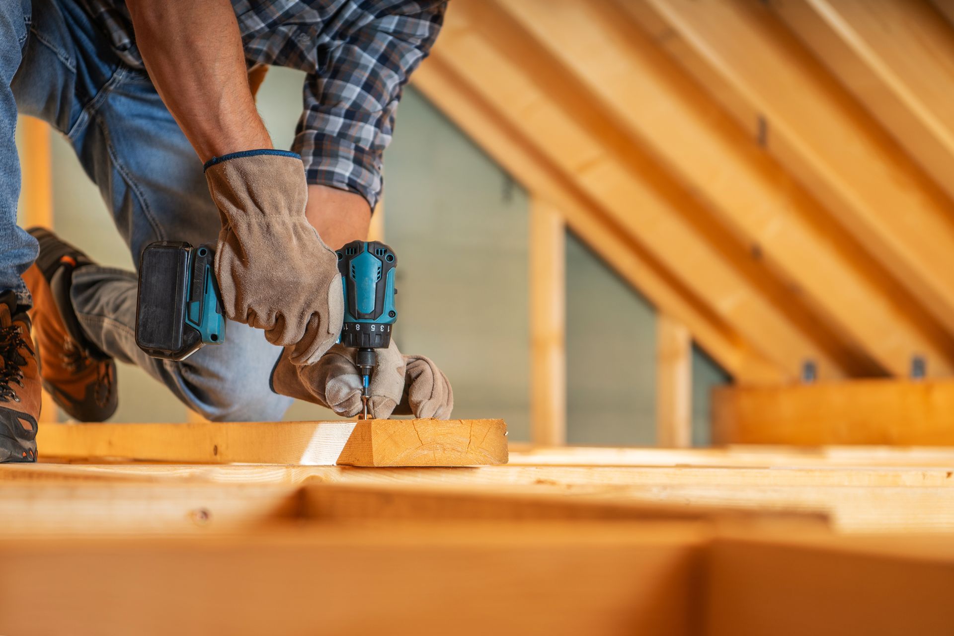 A person wearing work gloves uses a power drill on a wooden beam in an unfinished attic space. A person wearing work gloves uses a power drill on a wooden beam in an unfinished attic space.