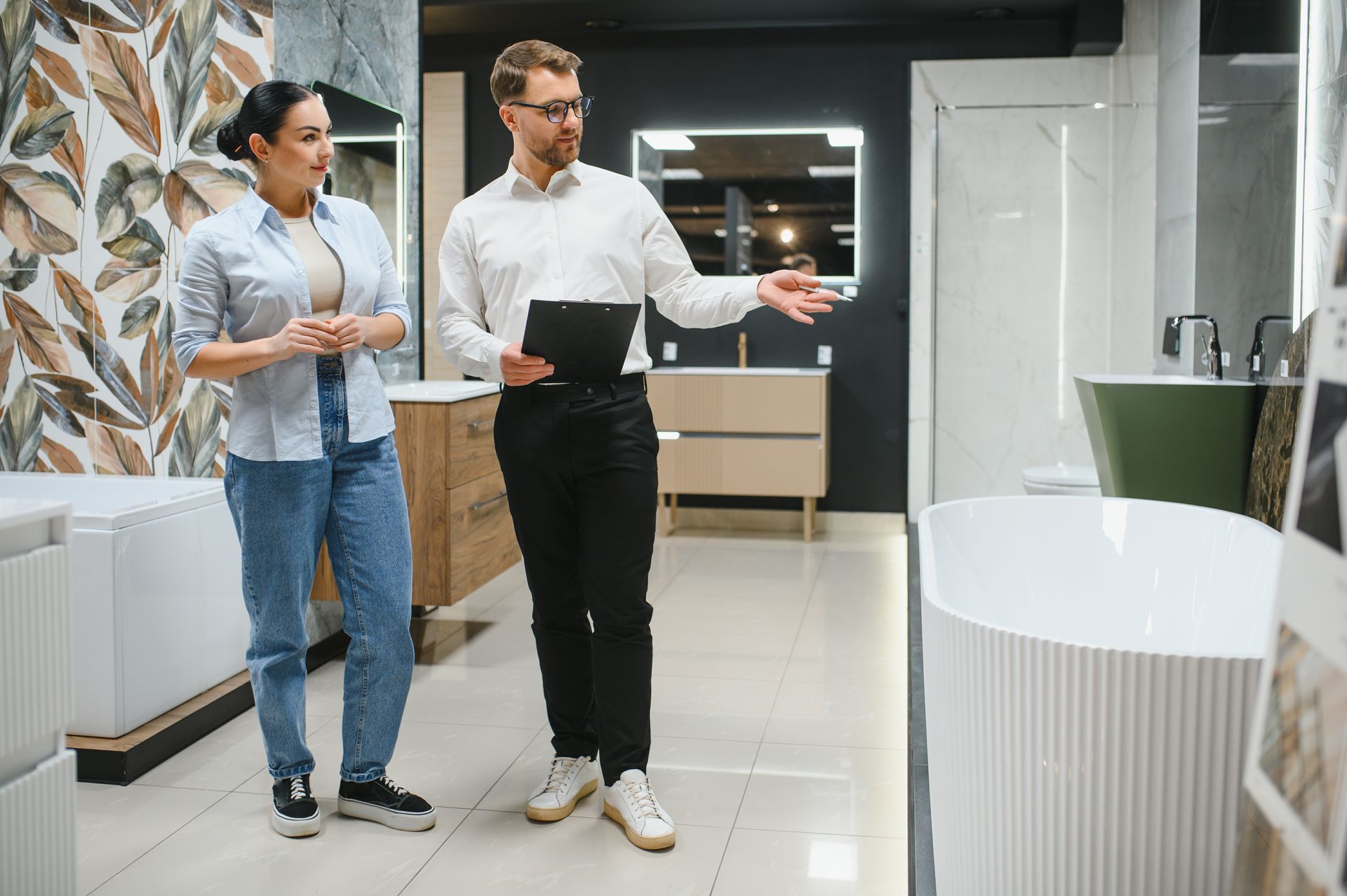 A sales representative holding a clipboard gestures toward a bathtub in a showroom while consulting with a customer. A sales representative holding a clipboard gestures toward a bathtub in a showroom while consulting with a customer.