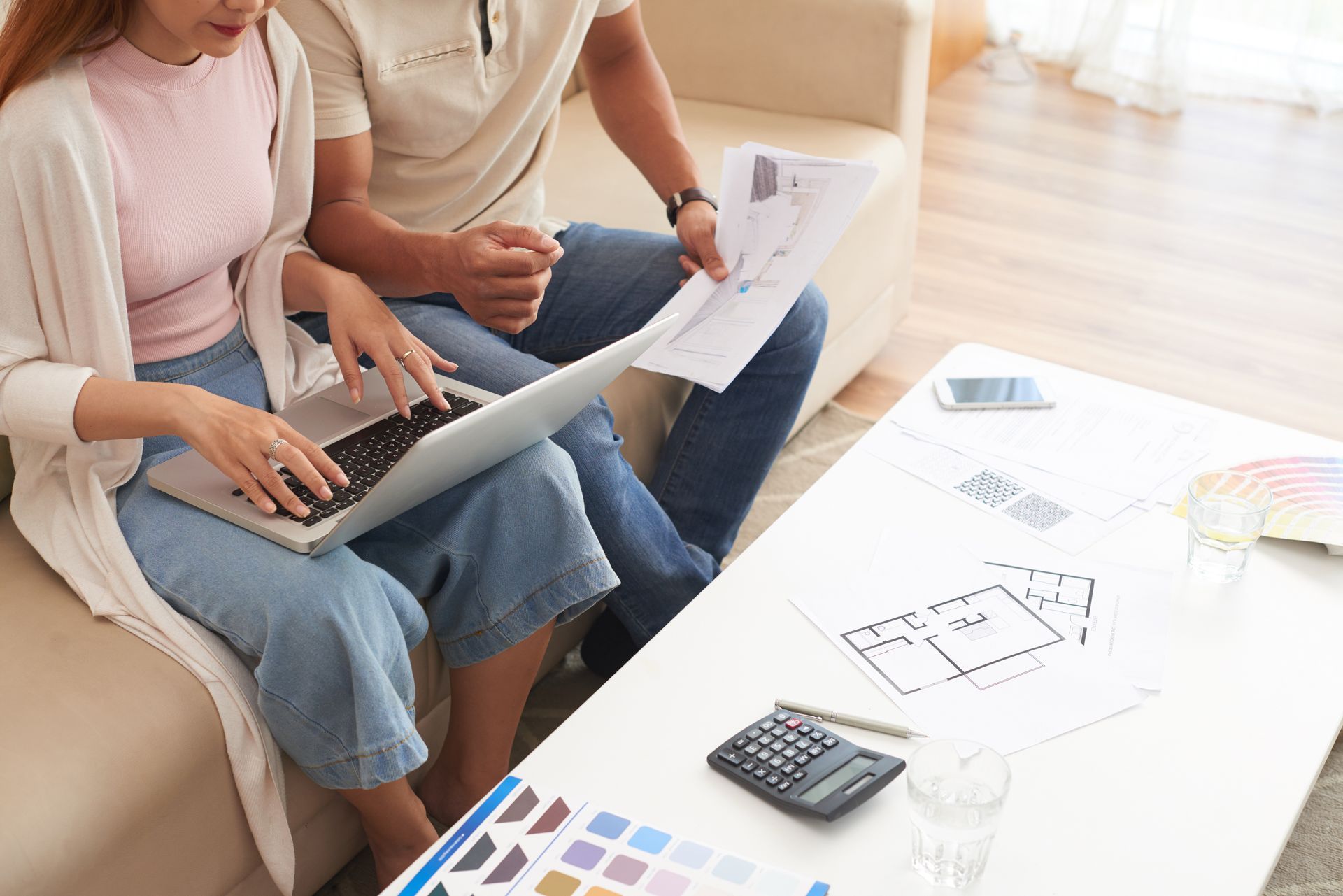 Two people sit on a sofa reviewing blueprints and using a laptop with a calculator and color swatches on a coffee table.