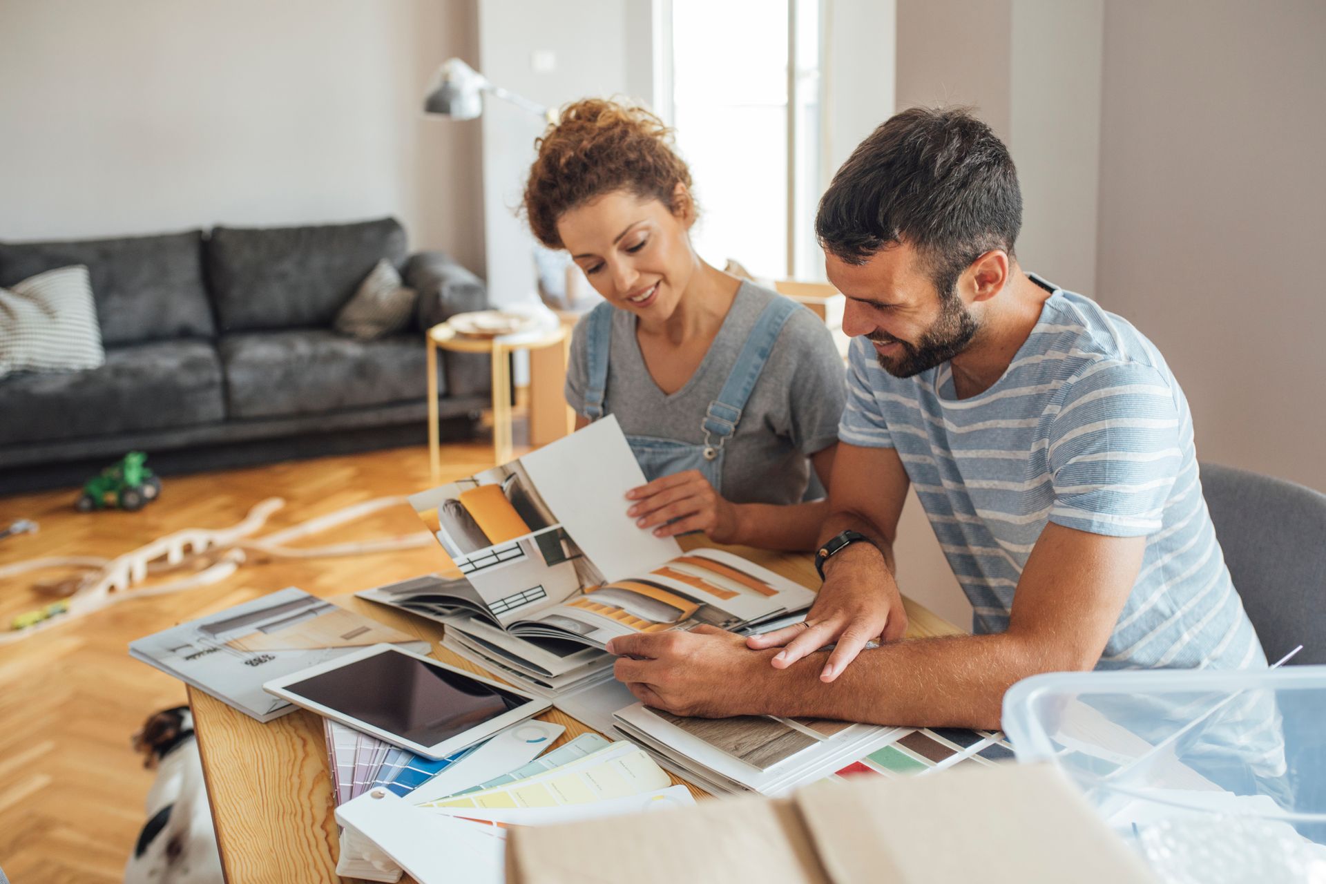 A couple sits at a table in a living room, reviewing fabric swatches and decor samples together while smiling. A couple sits at a table in a living room, reviewing fabric swatches and decor samples together while smiling.