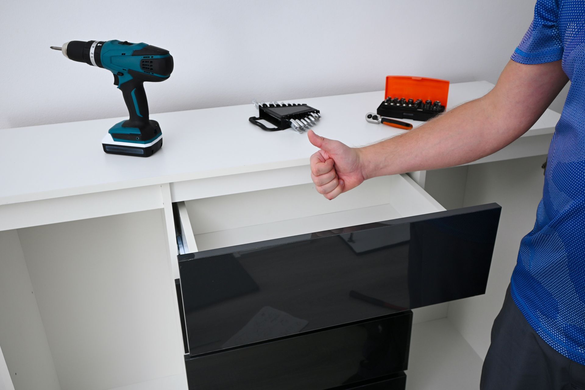 A person giving a thumbs-up next to an assembled white dresser with glossy black drawers and a power drill on top. A person giving a thumbs-up next to an assembled white dresser with glossy black drawers and a power drill on top.