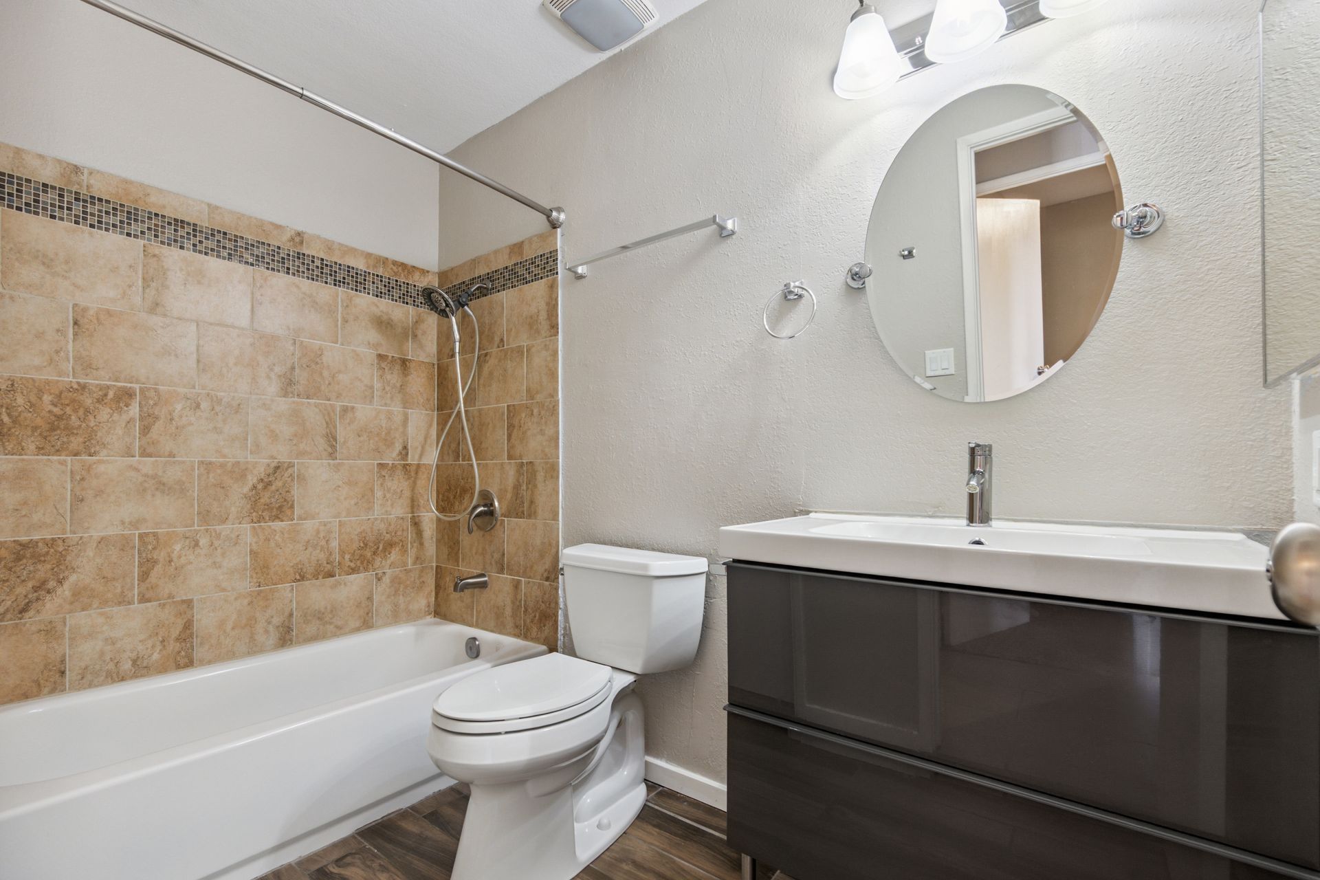 A bathroom with a beige tiled shower, white toilet, and a dark brown vanity with a circular mirror and light fixture. A bathroom with a beige tiled shower, white toilet, and a dark brown vanity with a circular mirror and light fixture.