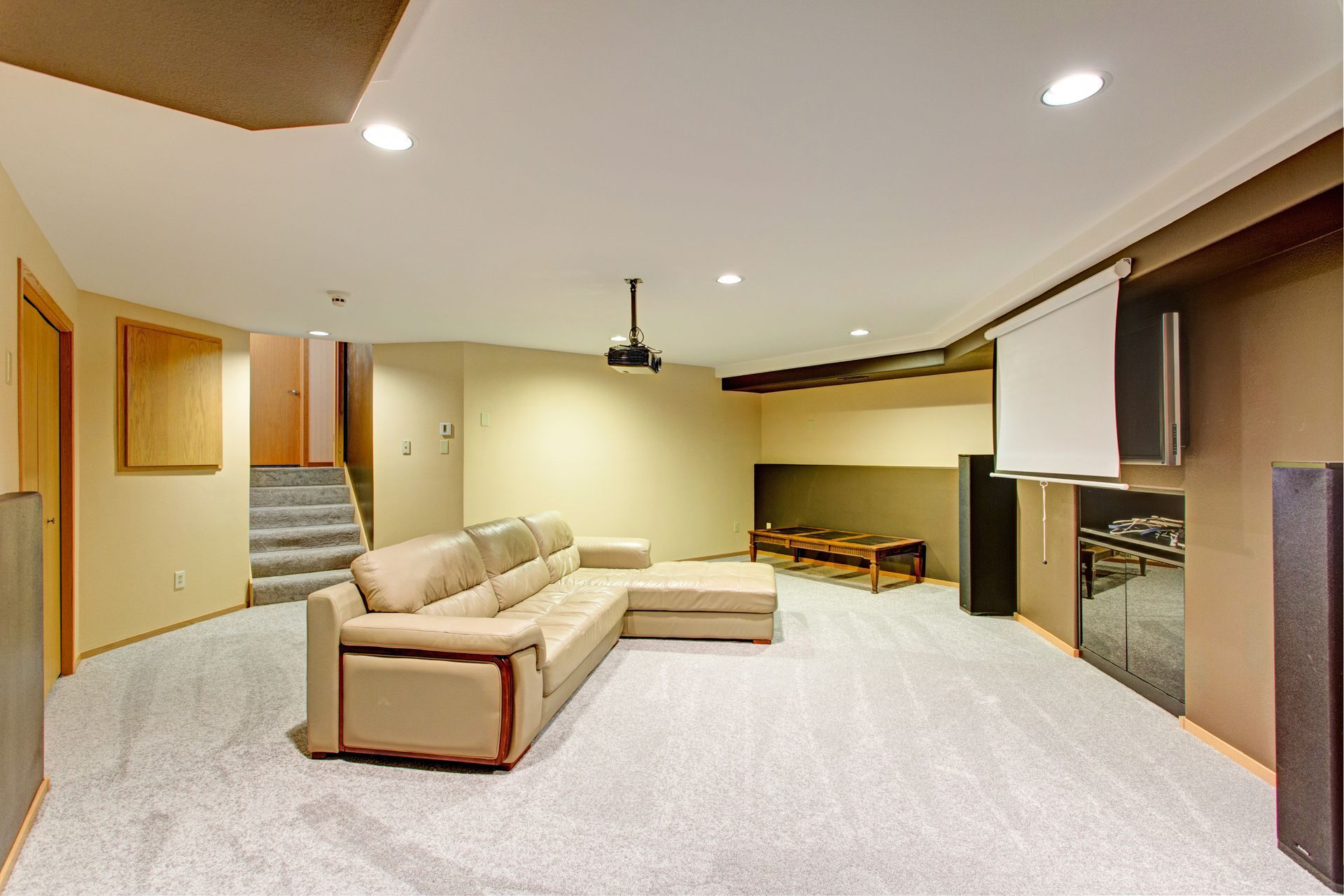 A basement home theater with a beige sectional sofa, projector, retractable screen, and gray carpet. A basement home theater with a beige sectional sofa, projector, retractable screen, and gray carpet.