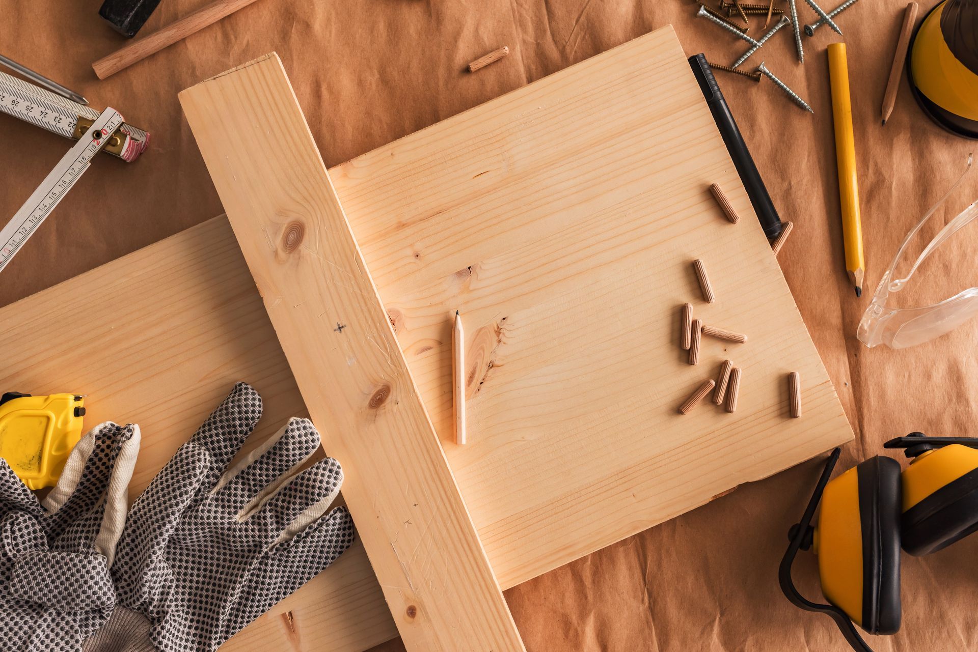 Top-down view of wood planks on a brown surface with a carpenter's square, wooden dowels, gloves, and safety gear. Top-down view of wood planks on a brown surface with a carpenter's square, wooden dowels, gloves, and safety gear.