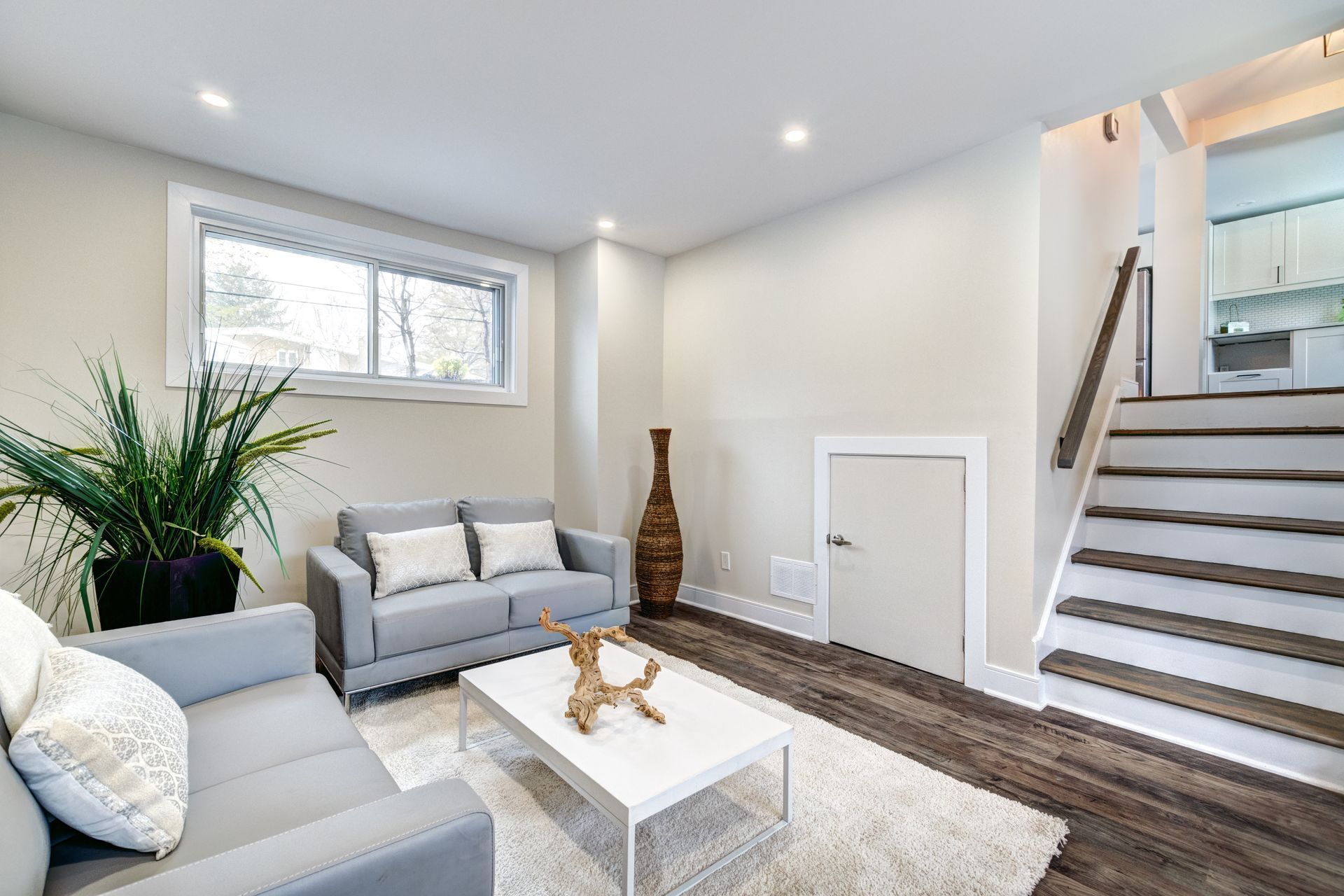 Bright living room with gray sofas, white coffee table, staircase, and natural light from a window