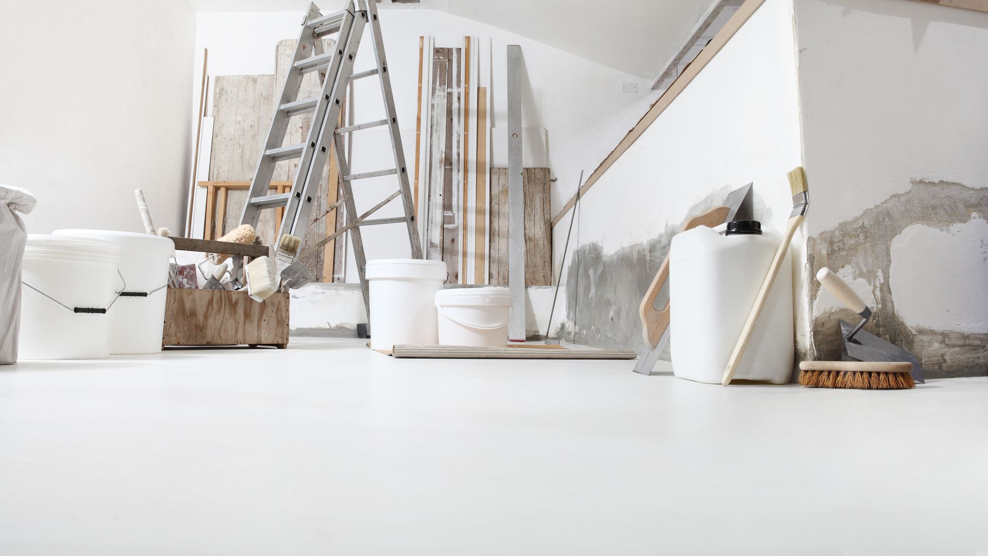 A ladder, paint buckets, and tools in a room under renovation with white walls and a smooth floor. A ladder, paint buckets, and tools in a room under renovation with white walls and a smooth floor.