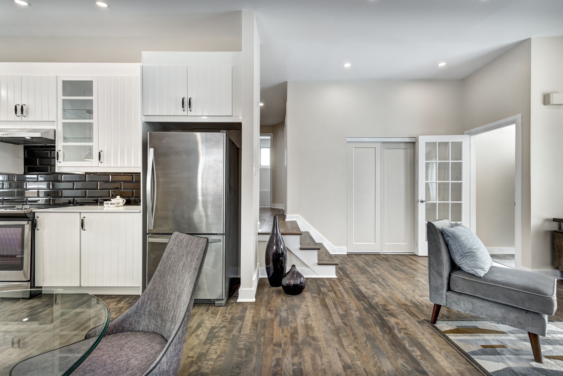 An open kitchen and living area featuring white cabinets, stainless steel appliances, wood floors, and gray armchairs. An open kitchen and living area featuring white cabinets, stainless steel appliances, wood floors, and gray armchairs.