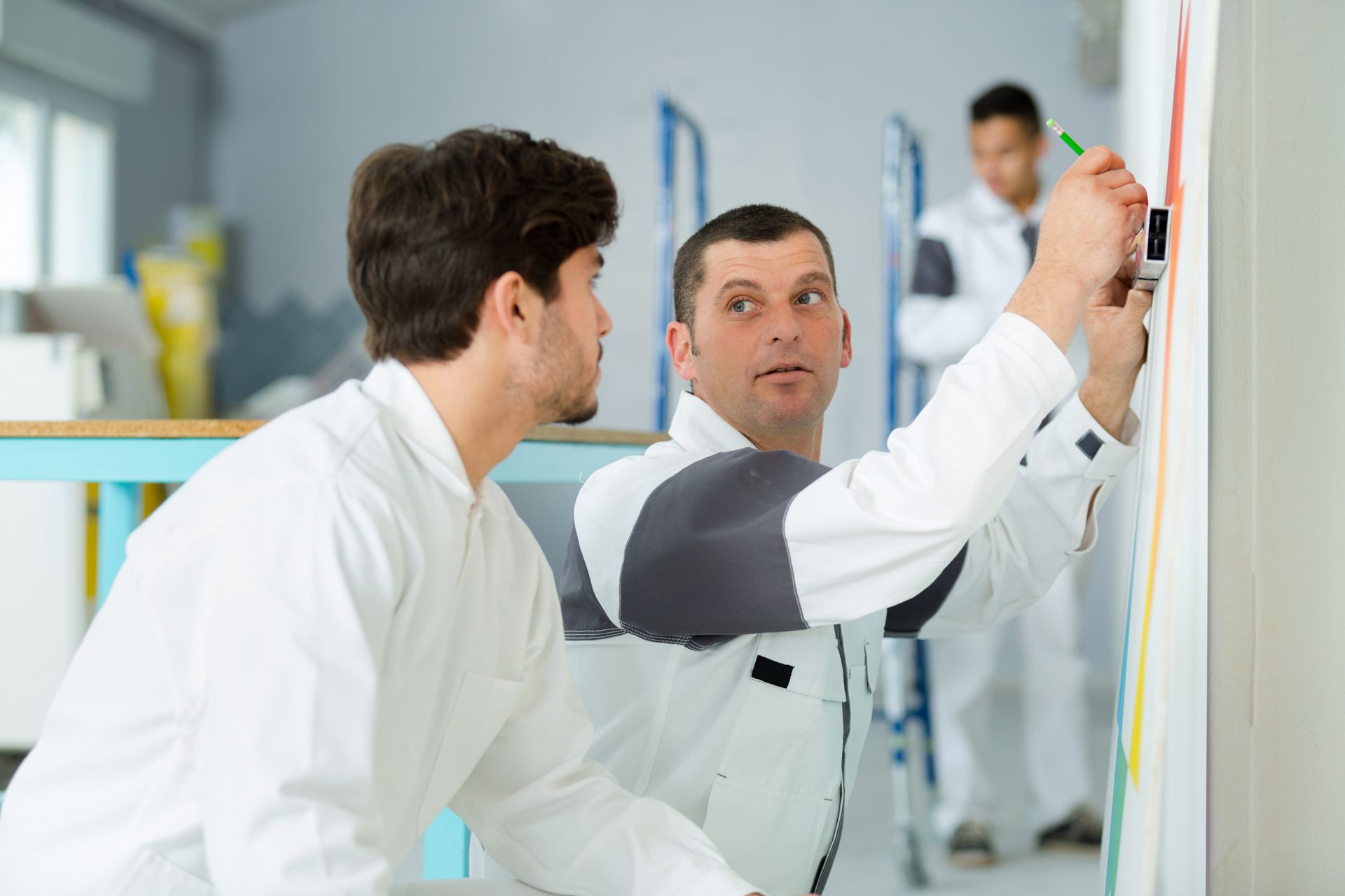 Two technicians in white uniforms working on a project, with one marking a measurement on a wall and another observing. Two technicians in white uniforms working on a project, with one marking a measurement on a wall and another observing.