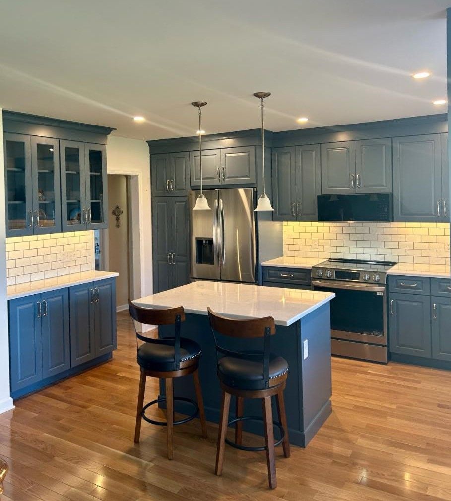 A modern kitchen with dark blue cabinets, white countertops, a center island with two bar stools, and hardwood floors. A modern kitchen with dark blue cabinets, white countertops, a center island with two bar stools, and hardwood floors.