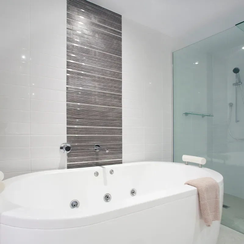 A white bathtub with jets sits in a modern bathroom with white walls and a decorative gray textured tile feature wall. A white bathtub with jets sits in a modern bathroom with white walls and a decorative gray textured tile feature wall.