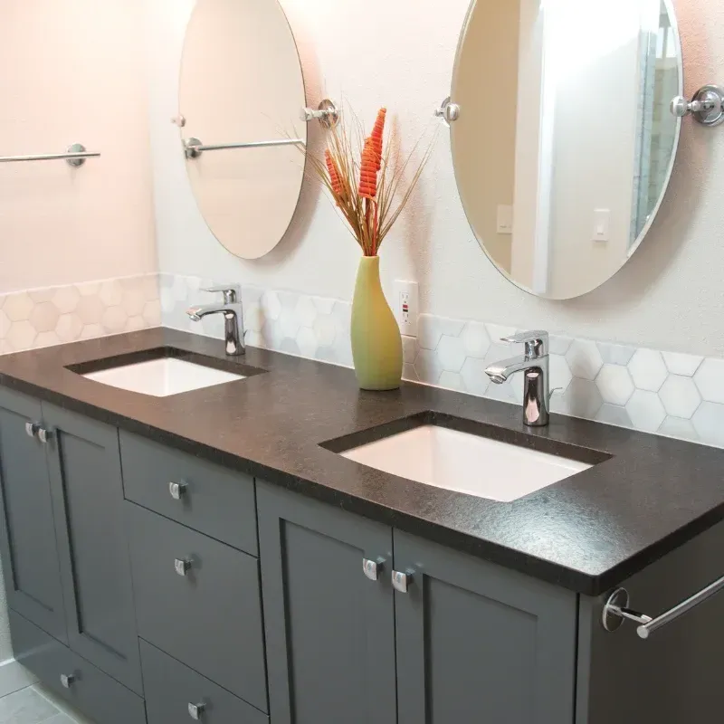 Double vanity with a dark countertop, grey cabinets, two oval mirrors, and white hexagonal tile backsplash. Double vanity with a dark countertop, grey cabinets, two oval mirrors, and white hexagonal tile backsplash.