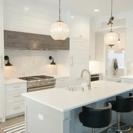 A bright, modern kitchen featuring a white island with black stools, white cabinets, and a grey wood range hood. A bright, modern kitchen featuring a white island with black stools, white cabinets, and a grey wood range hood.